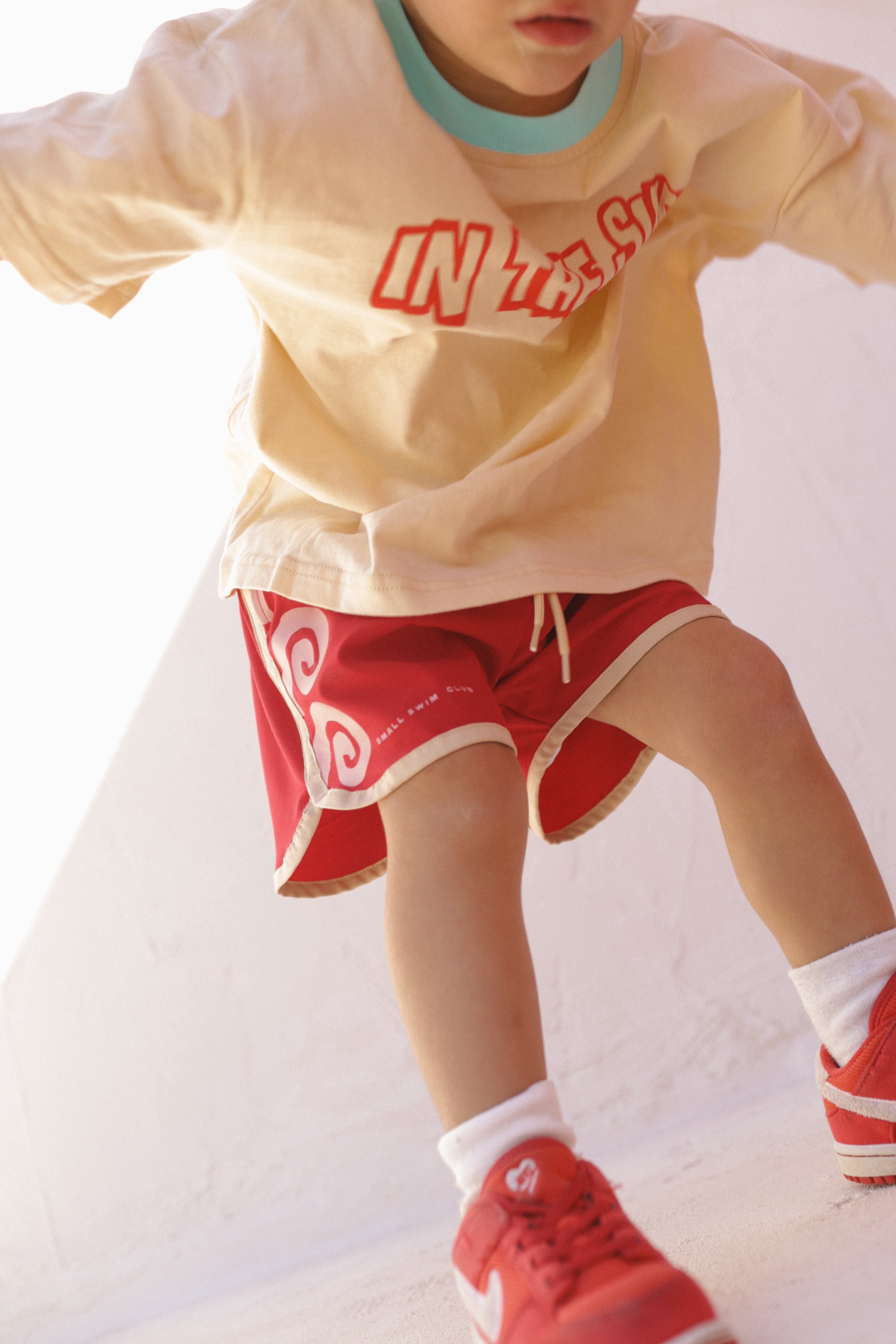 A child is in motion, wearing a cream T-shirt with red lettering, Maroon Swirl Boardshorts by SMALL SWIM CLUB featuring a swirl graphic and elastic drawstring waistband, white socks, and red sneakers. The focus is on the vivid clothing against a plain background. Face is out of frame.