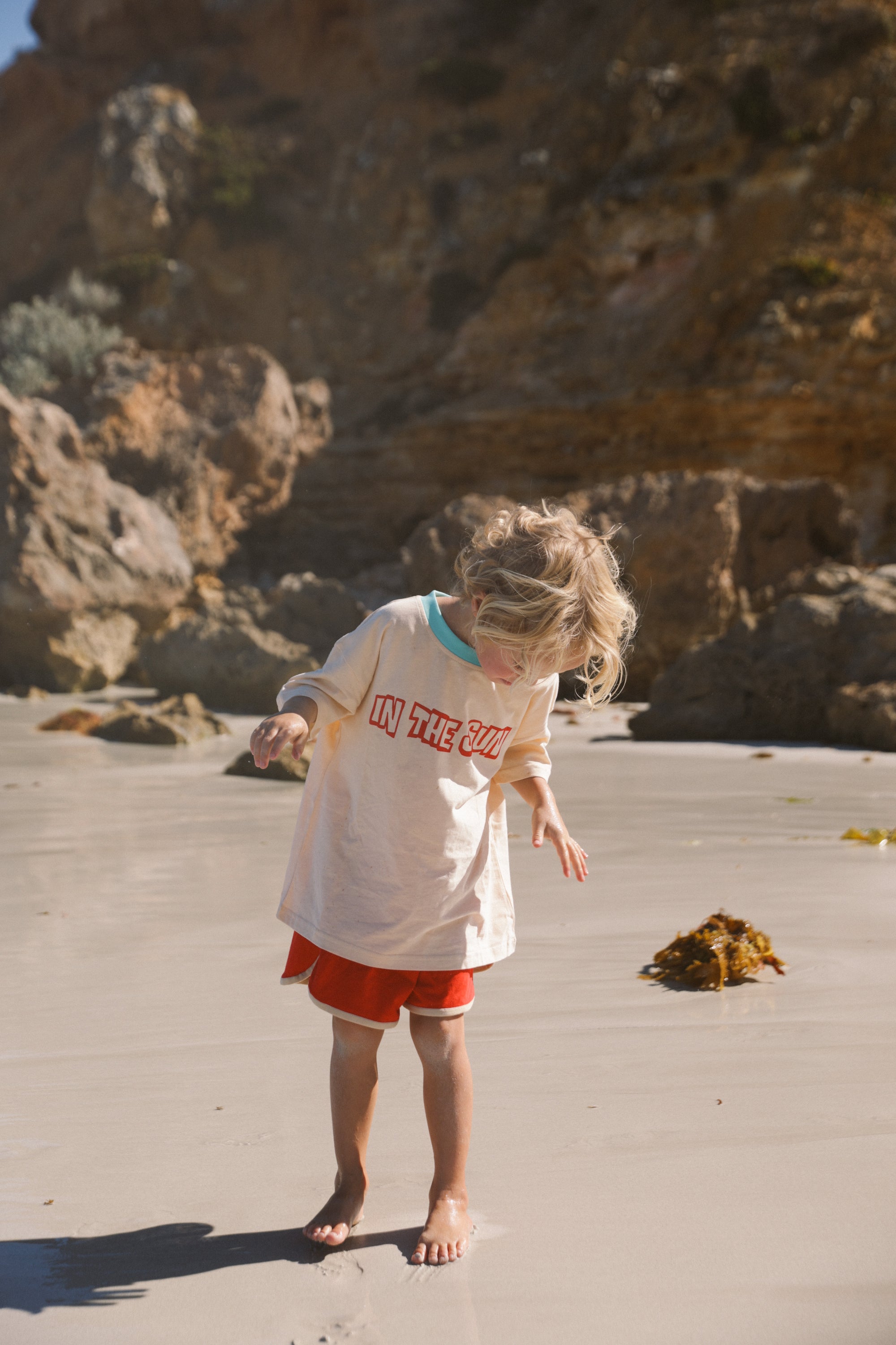 A child with blonde hair explores a sandy beach in red shorts and an oversized "In the Sun Tee Cream" by SMALL SWIM CLUB. The scene is framed by rocky cliffs and seaweed under a clear sky.