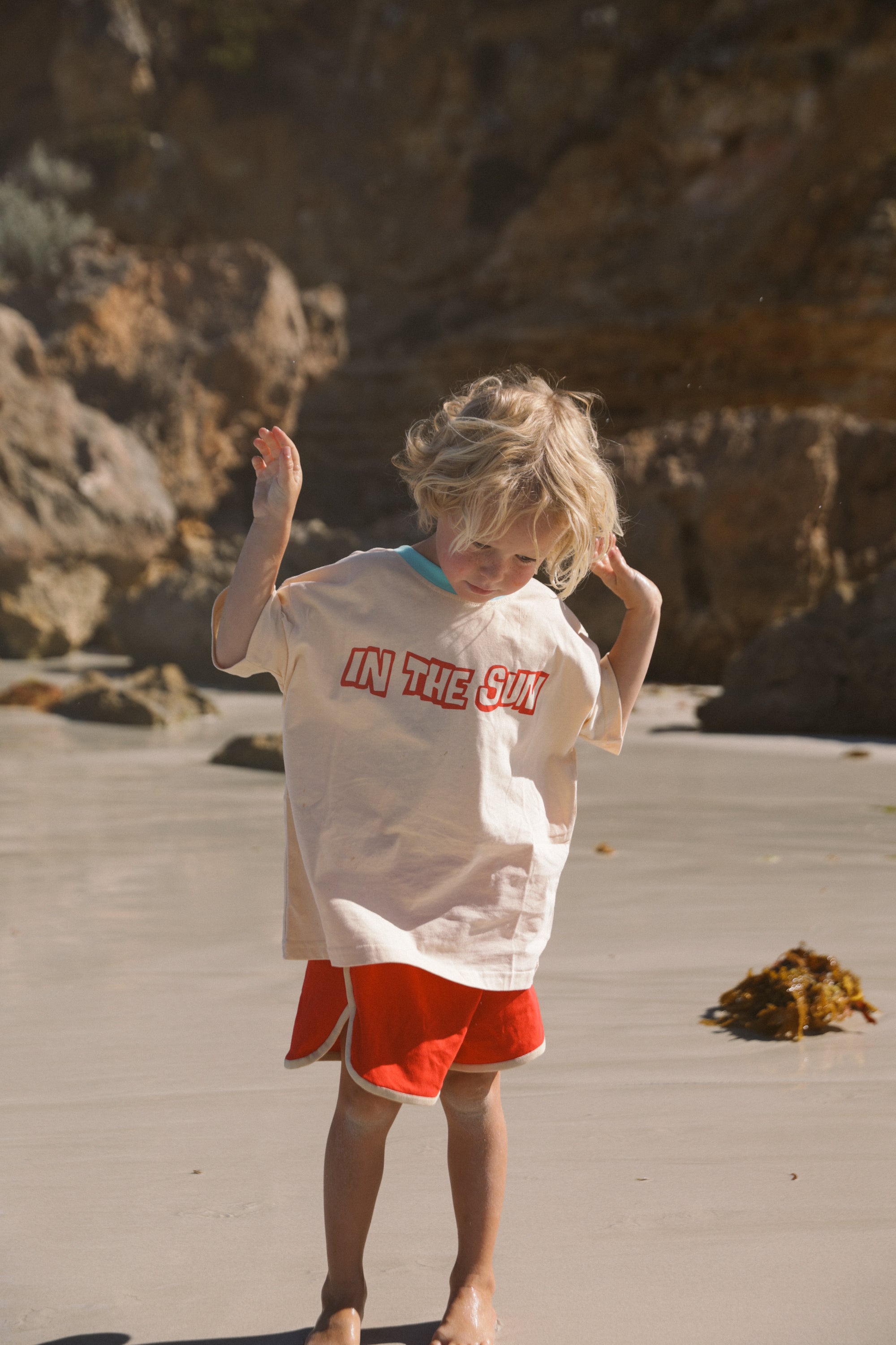 A child with blond hair stands on a sandy beach in SMALL SWIM CLUB’s In the Sun Tee Cream and orange shorts, hands raised and gazing at the sand, with rocky formations in the background.