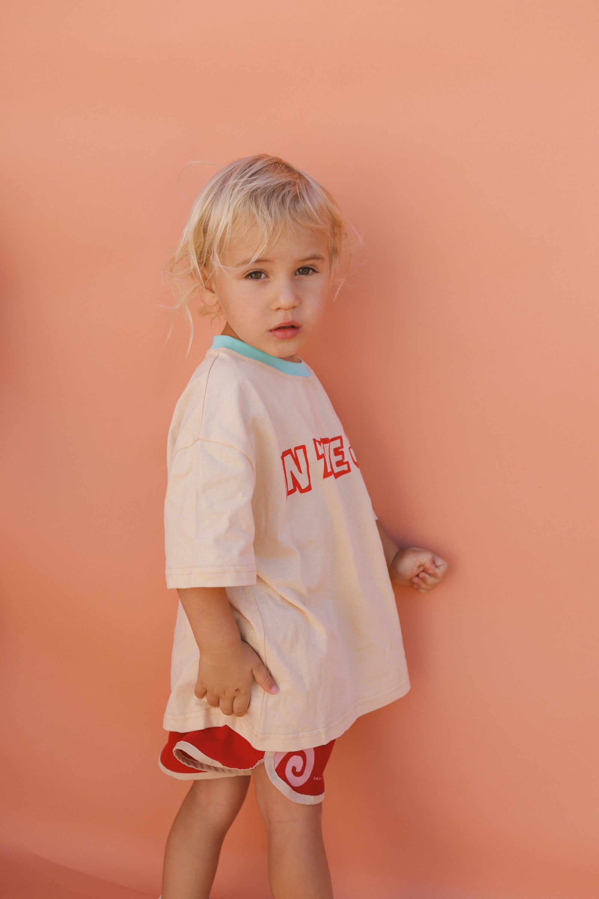 A young child with blonde hair poses against a peach backdrop, wearing the In the Sun Tee Cream by SMALL SWIM CLUB in size 7-8, along with red shorts. The graphic tee's loose fit complements their neutral expression as they gaze at the camera.