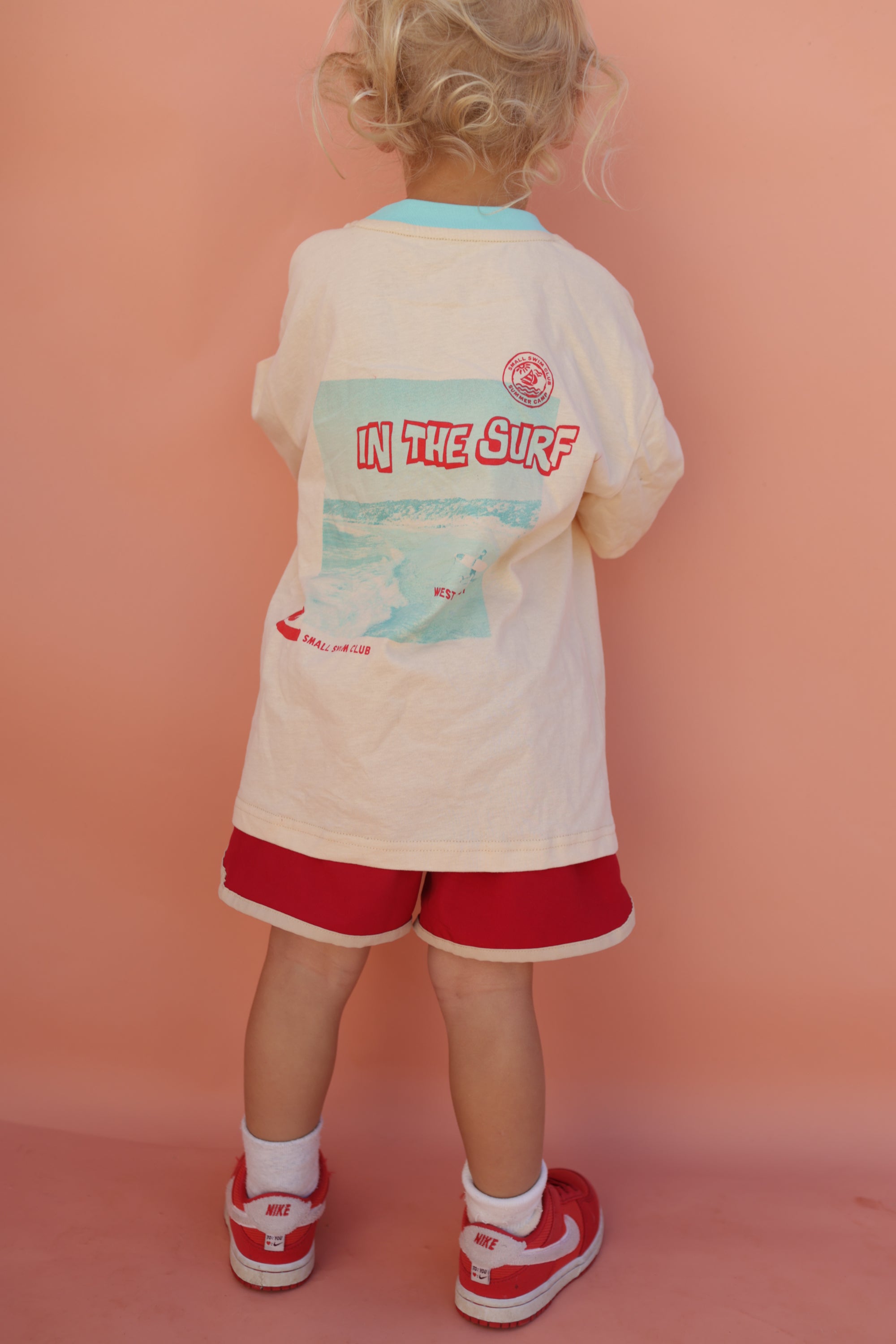 A child with curly blonde hair faces a peach-colored wall, wearing the SMALL SWIM CLUB's In the Sun Tee Cream, featuring a vibrant graphic, paired with red shorts. The look is completed with red Nike sneakers and white socks for a sporty flair.