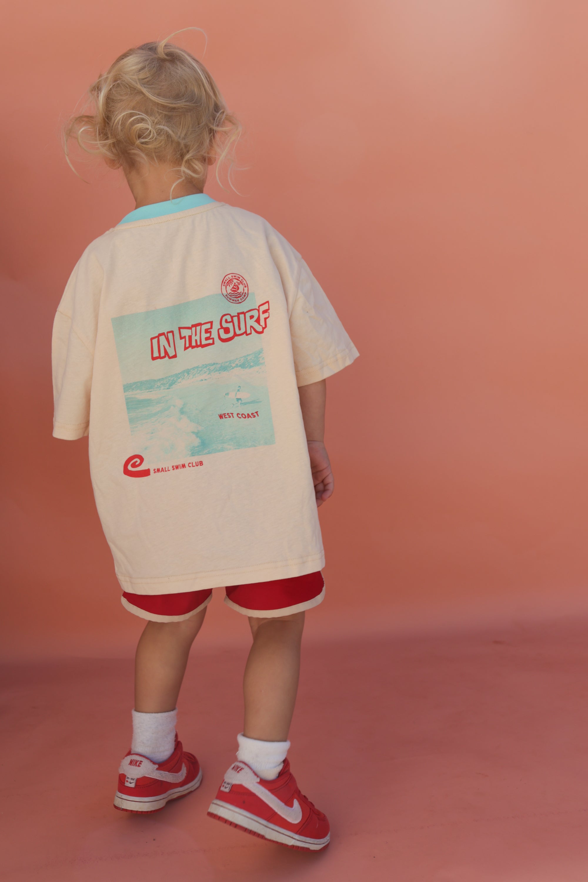 A blonde child stands against a peach background, wearing the SMALL SWIM CLUB's oversized "In the Sun Tee Cream" with a beach-themed graphic and "IN THE SURF" on the back, paired with red shorts and matching sneakers.