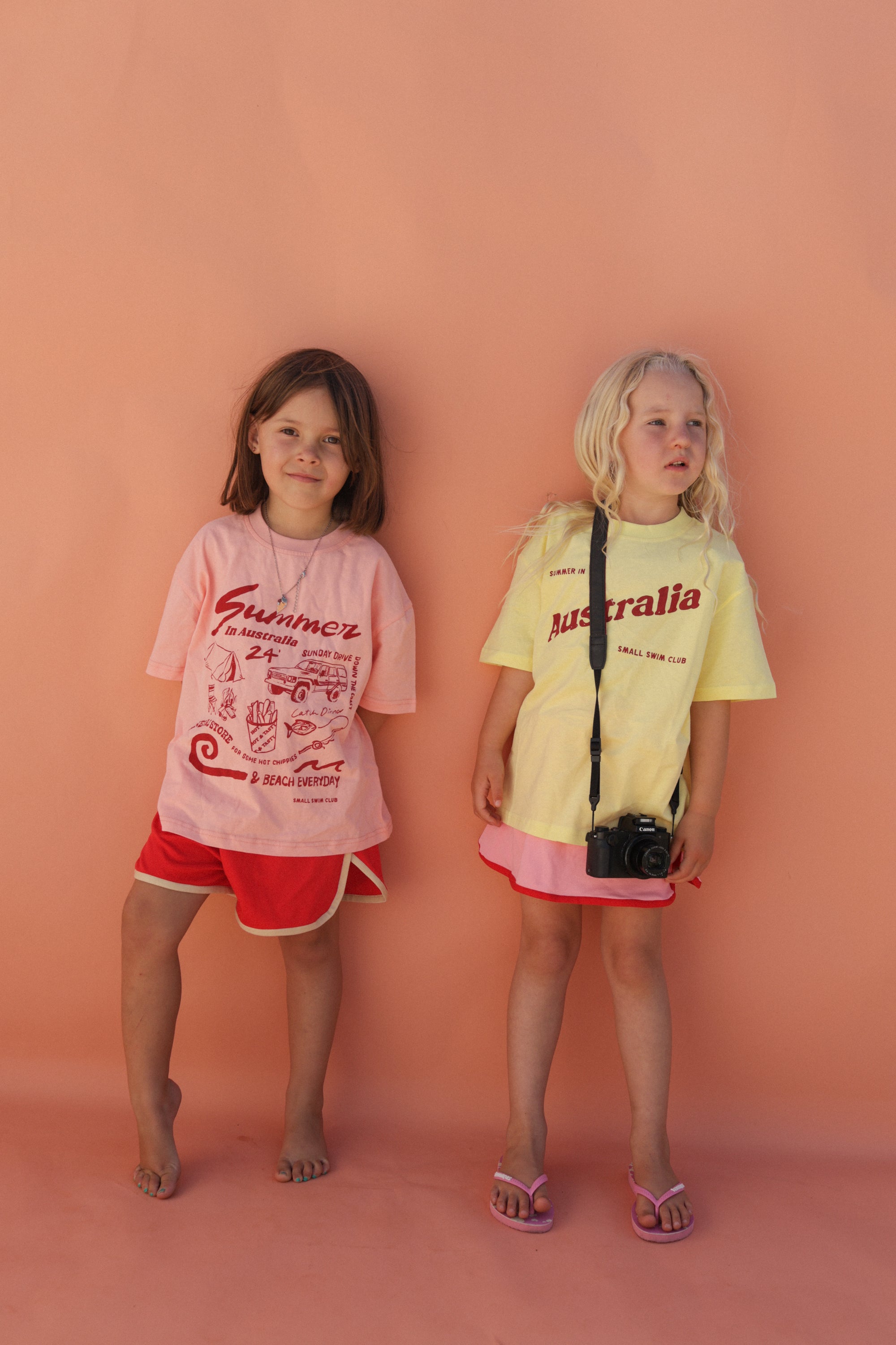 Two young children stand against a peach background. One wears a pink shirt with maroon "Summer" text and red shorts, while the other sports the SMALL SWIM CLUB's "Summer in Australia Tee Lemon," perfect for an Aussie summer, and holds a camera. Both appear relaxed and cheerful.