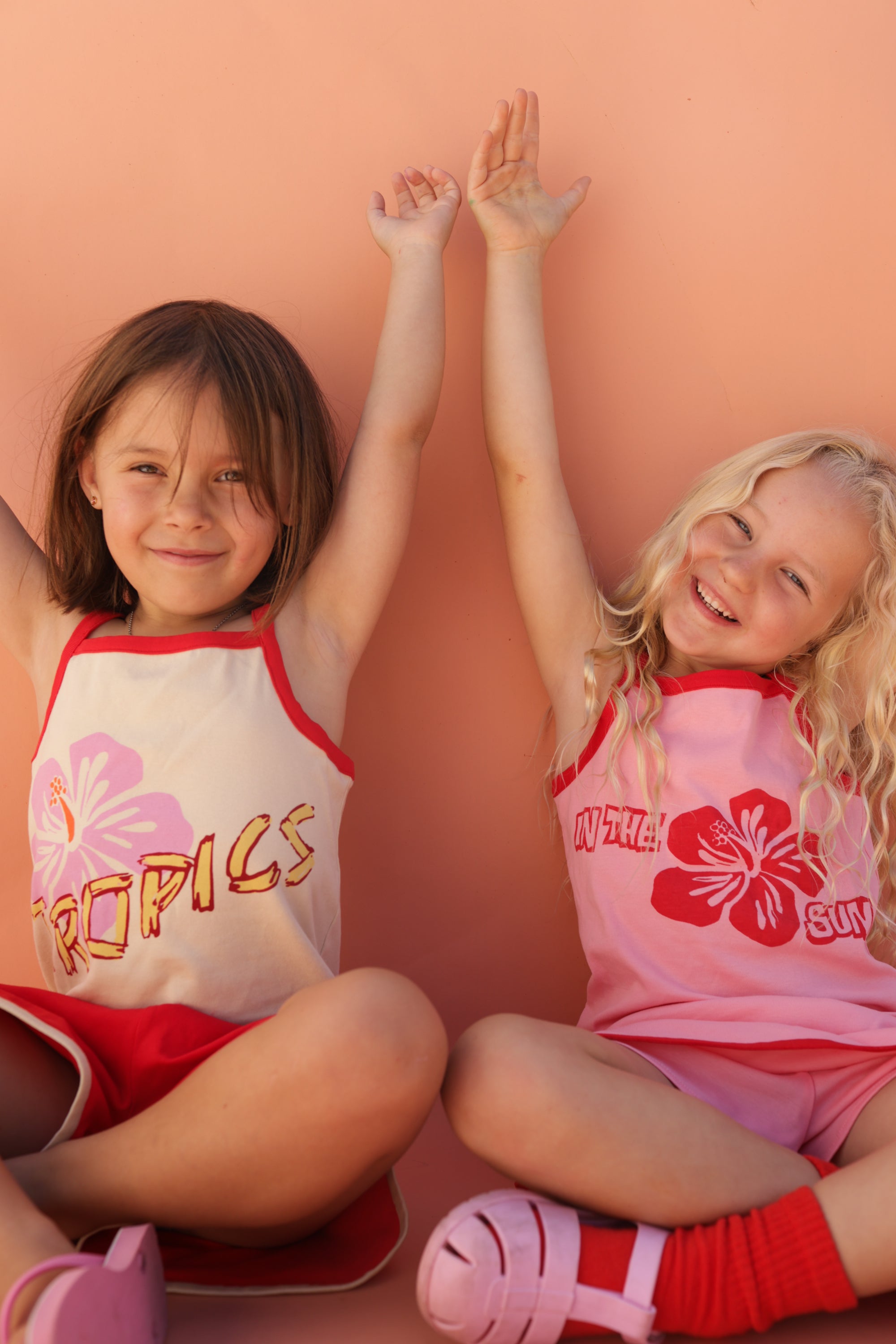 Two young girls sit against a peach wall, smiling with arms raised. One with brown hair wears the SMALL SWIM CLUB "Tropics" shirt and shorts, while the blonde wears a SMALL SWIM CLUB Hibiscus Singlet Pink/Red and a pink skort. Both wear sandals, exuding joy and playfulness.