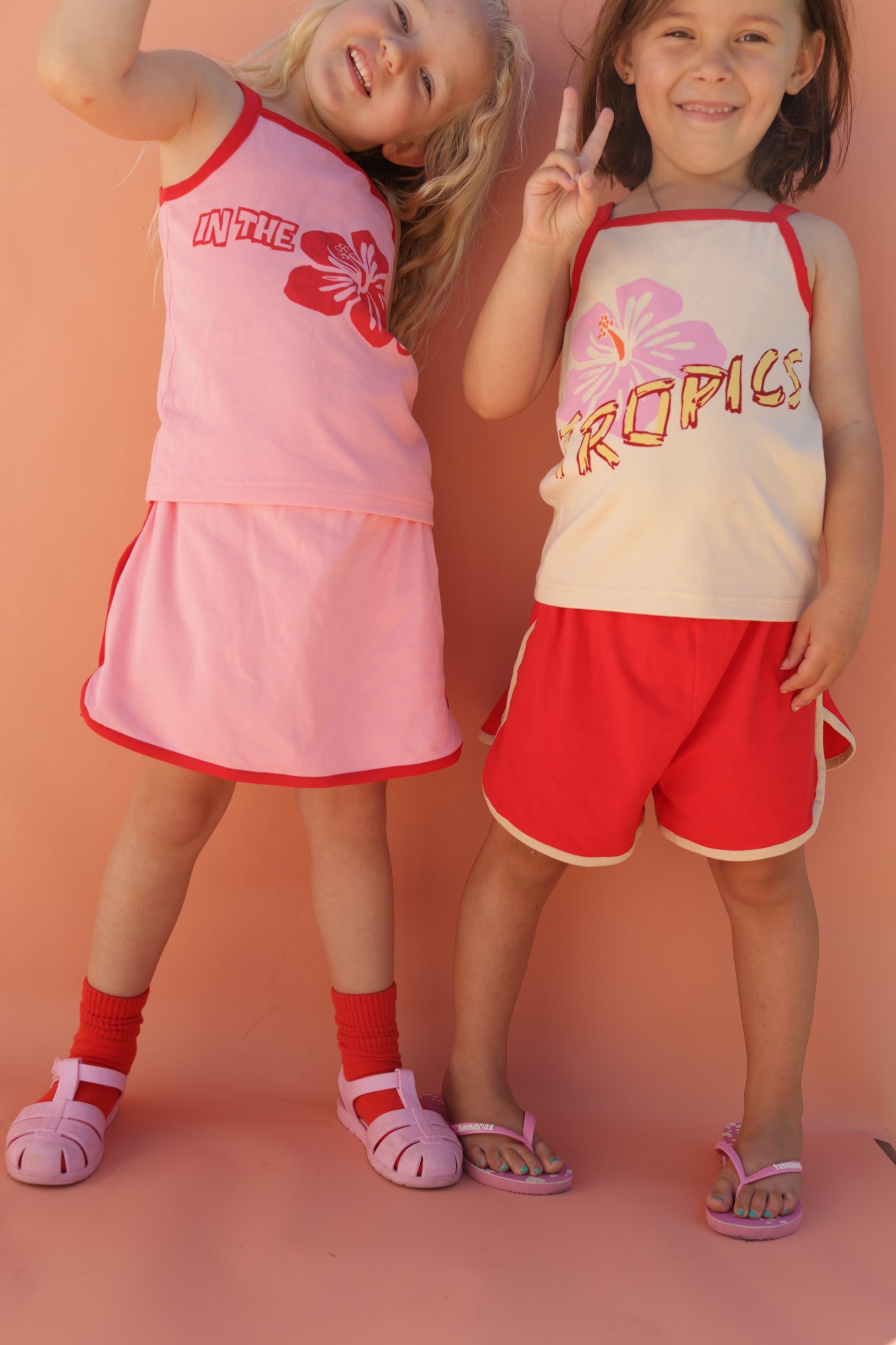 Two young children pose and smile against a peach background in coordinating summer outfits by SMALL SWIM CLUB: a pink floral skirt and 70's Skort Pink/Red in size 7-8. One child shows a peace sign, both wearing sandals. All items are made of 100% cotton.