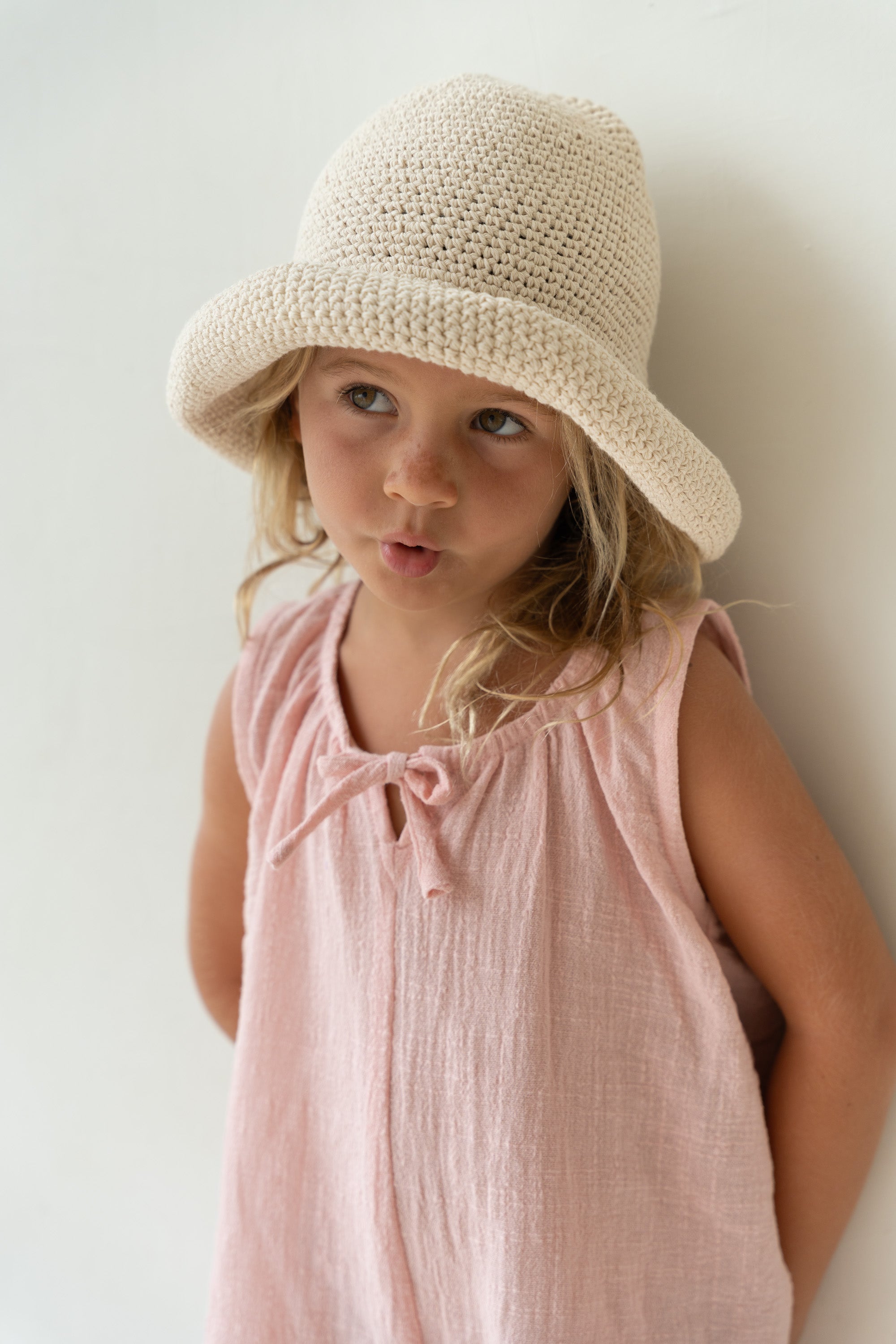 A young child with long hair wears the Crochet Hat Ecru by ILLOURA THE LABEL and a sleeveless pink dress. They stand against a light-colored background, making an expressive face with their lips pursed and eyes looking to the side.