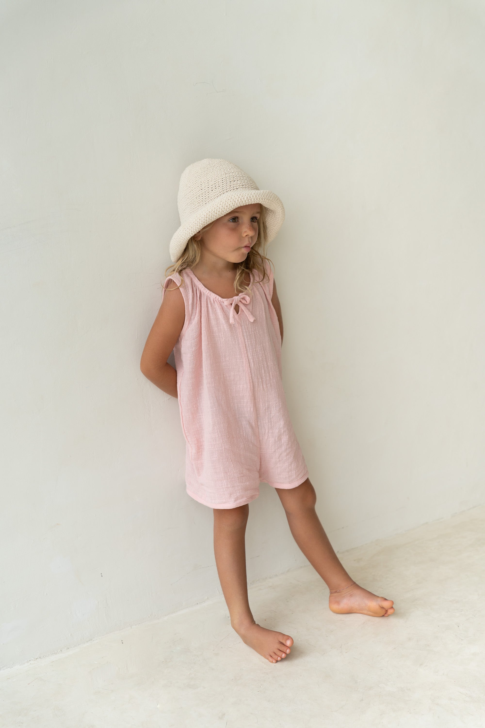 A young girl stands against a light-colored wall, wearing a light pink dress and the Crochet Hat Ecru from ILLOURA THE LABEL. She is barefoot and looking to the side with a relaxed posture, set in a simple and minimalistic environment.