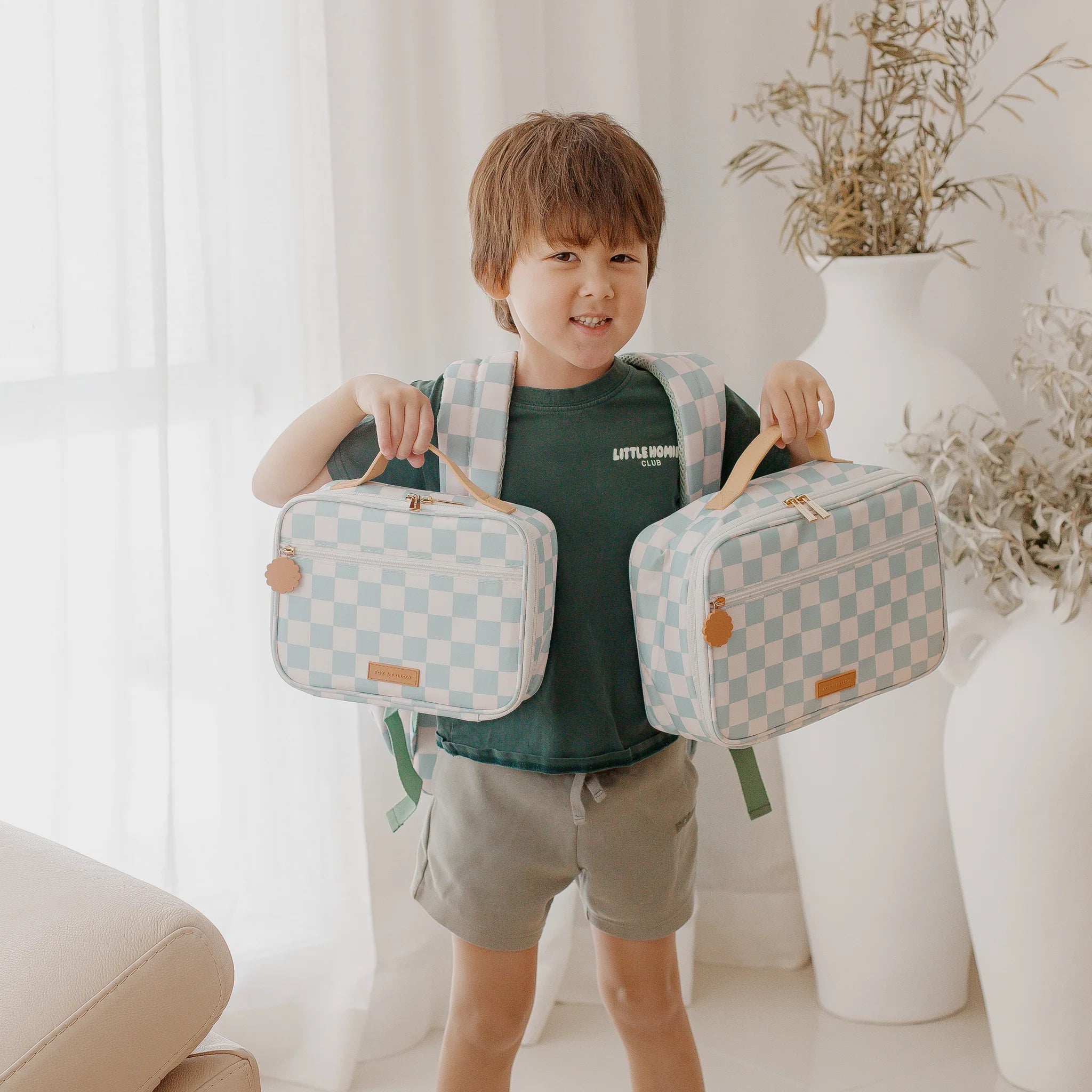 A young child in a green outfit stands indoors, smiling and holding two FOX & FALLOW Sage Check Large Lunch Bags, with a matching backpack on their back. Neutral decor and natural light fill the room.