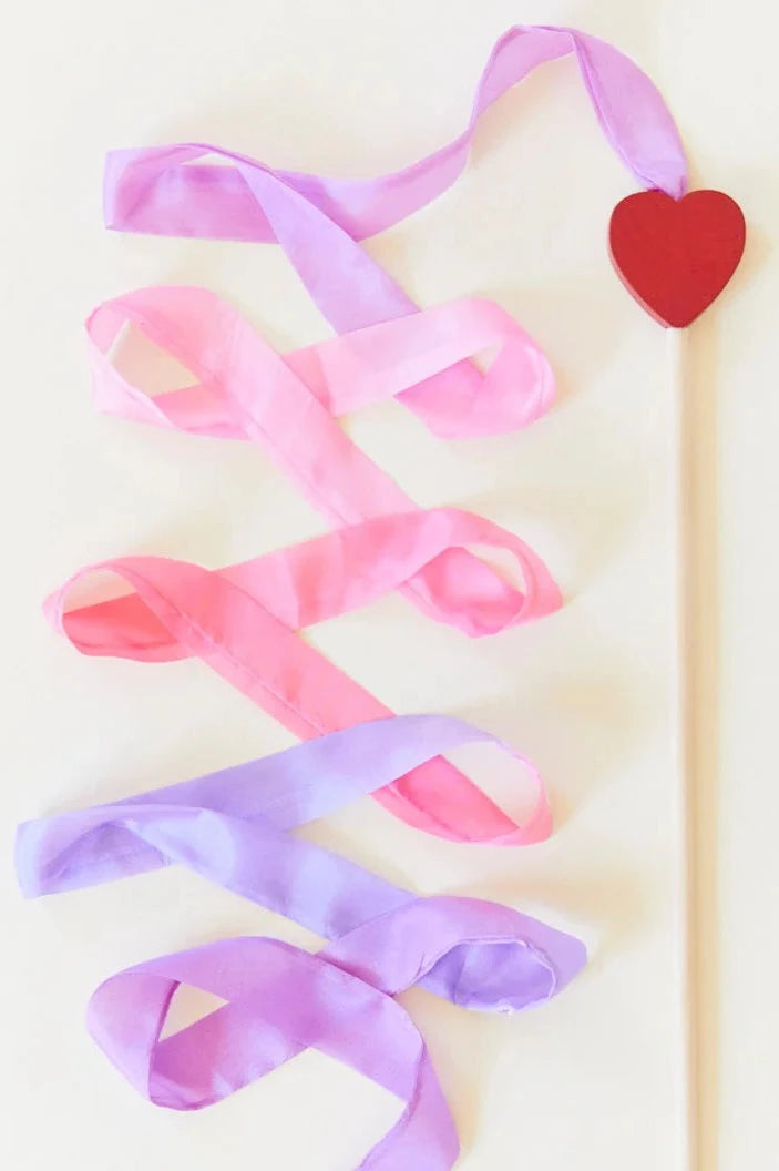 The Large Blossom Streamer by SARAH'S SILKS features a wooden wand topped with a red heart, adorned with intertwined pink and purple silk ribbons that create a crisscross pattern. These streamers cascade gracefully against a light background, crafted from sustainably sourced materials.