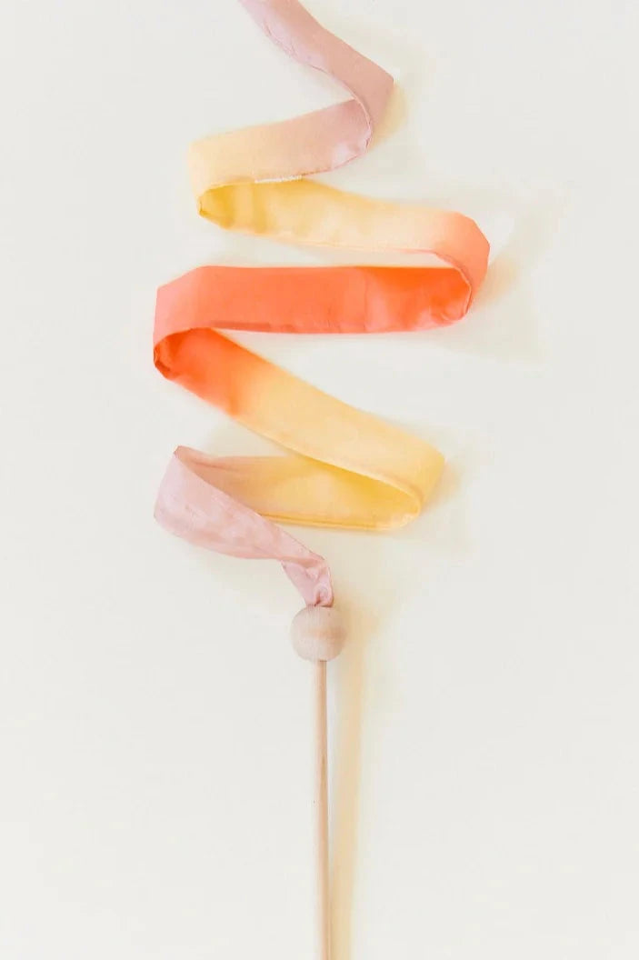 The Mini Desert Streamer by SARAH'S SILKS, crafted from sustainably sourced materials, is a wooden wand with a silk streamer showcasing a vibrant gradient that transitions from pink to orange to yellow. This elegant piece stands out against a plain white background.