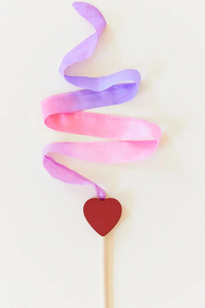 Introducing the Mini Blossom Streamer from SARAH'S SILKS, a charming wooden wand with vibrant purple and pink ribbons spiraling around a slender stick, accented by a red heart at its base. Crafted from sustainably sourced materials, it stands elegantly against a white background.