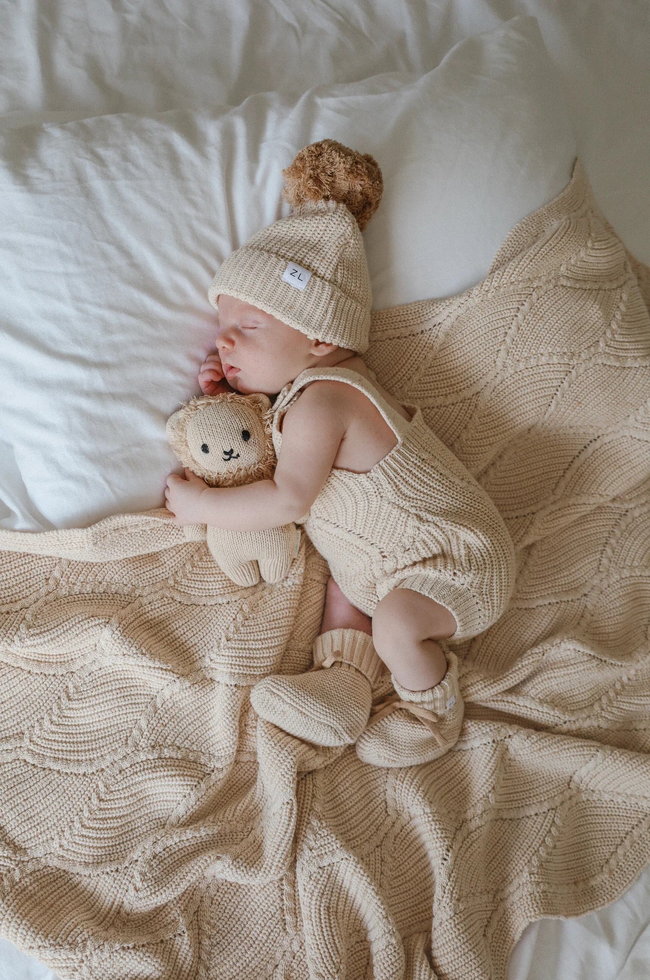 A baby sleeps peacefully on a knit blanket, wearing a matching ZIGGY LOU Beanie Biscotti Fleck, romper, and booties. The baby is cuddling a small knit stuffed animal. The blanket, clothing, and stuffed animal are all in 100% cotton biscotti fleck material, creating a cozy and warm scene.