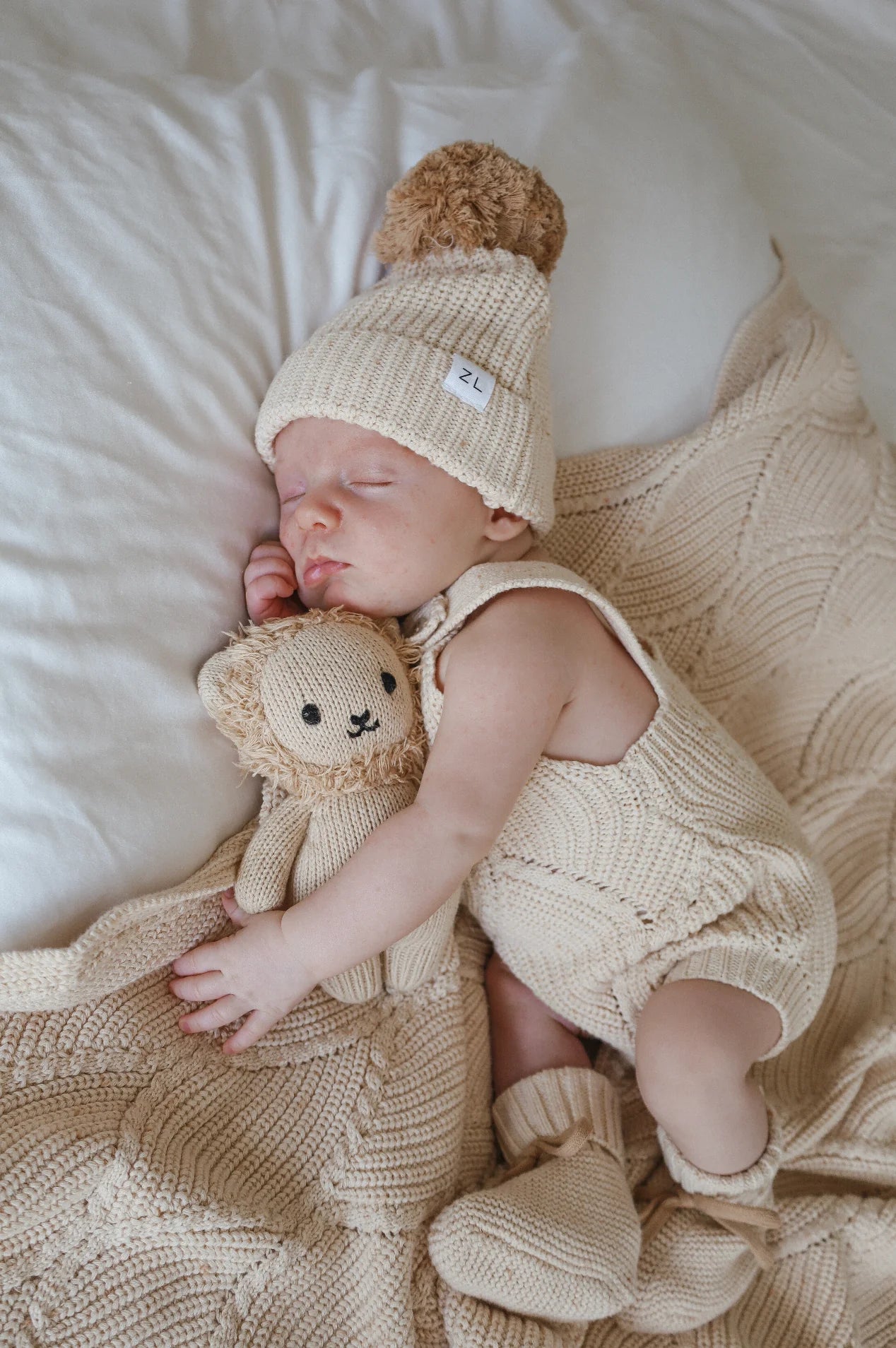 An infant is sleeping on a cream-colored knitted blanket. The baby, dressed in a light-colored, 100% cotton outfit with matching booties and a chunky knit Beanie Biscotti Fleck from ZIGGY LOU, is cuddling a small knitted lion toy while lying on their side.