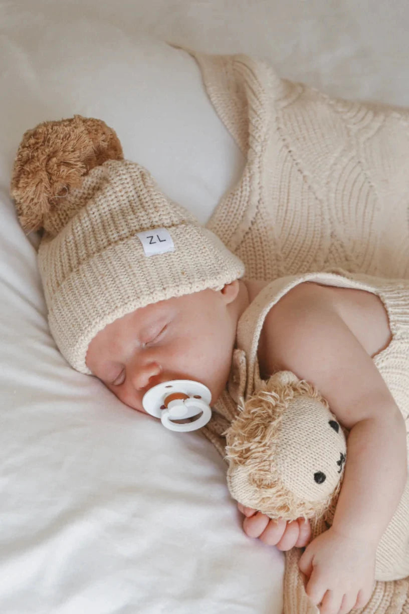 A sleeping baby dressed in a cozy beige knit outfit lies on a white bedsheet. The baby is wearing the Beanie Biscotti Fleck by ZIGGY LOU, featuring a chunky knit design with a fluffy pompom, and is holding a soft toy that resembles a teddy bear. The 100% cotton pacifier in their mouth completes the picture of serene slumber.