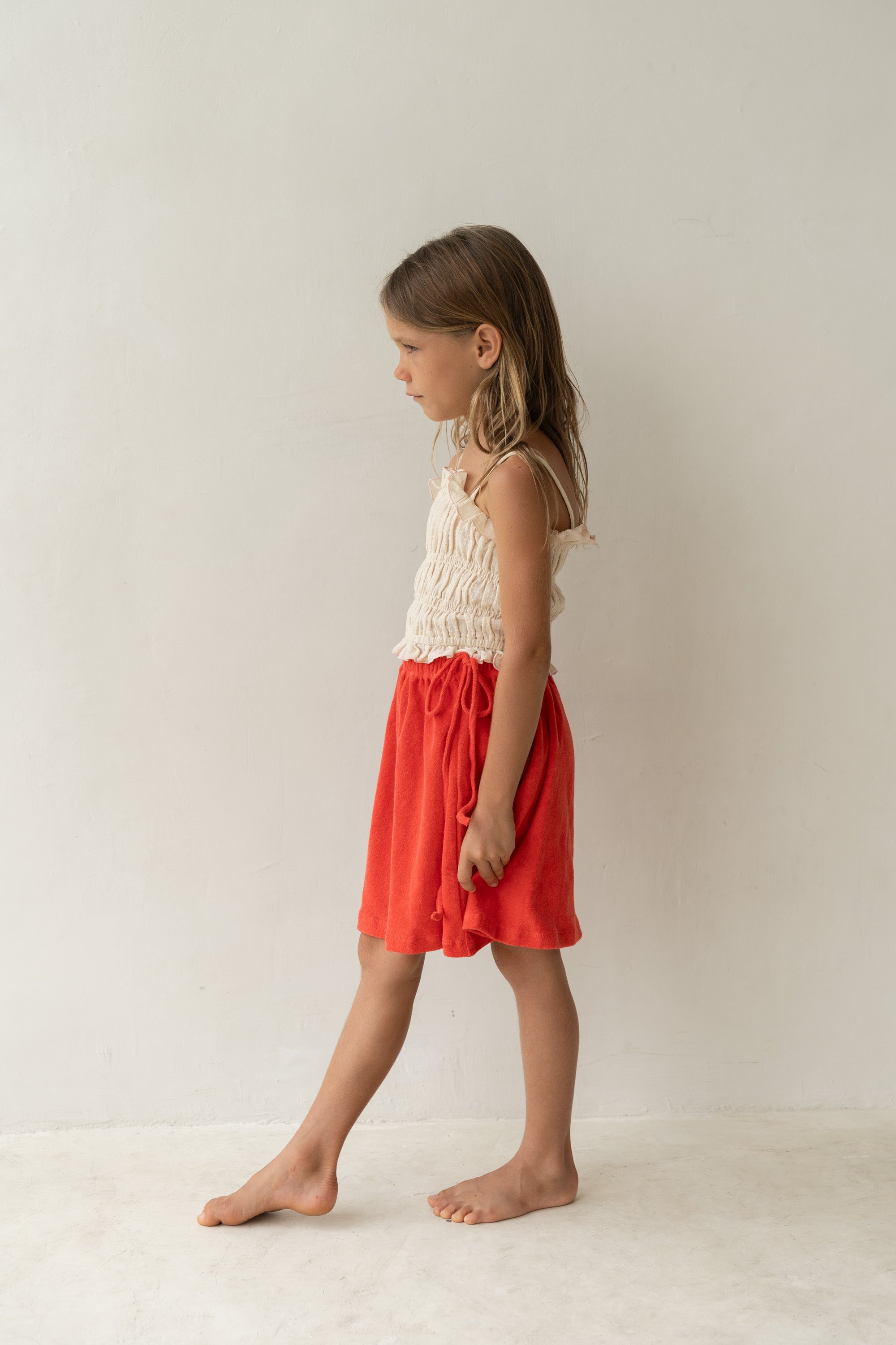 A young girl in a sleeveless cream top and the ILLOURA THE LABEL Lennie Skirt Watermelon with an elasticated waistband stands barefoot against a plain light-colored wall, looking to the side. Her hair is long and loose. The setting is minimalist and bright.