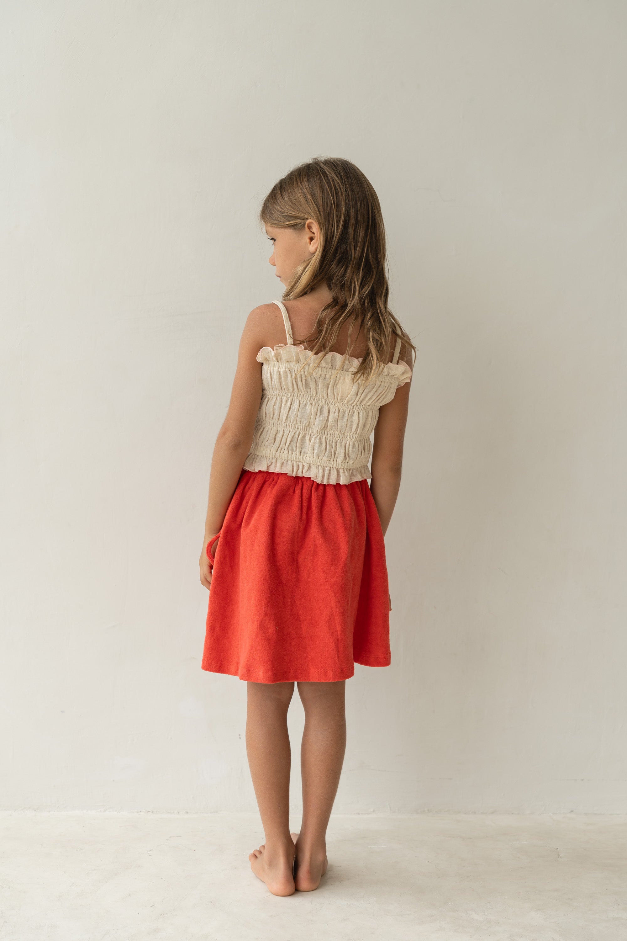 A young girl faces the wall, wearing a cream-colored smocked top and the Lennie Skirt Watermelon by ILLOURA THE LABEL, which features an elasticated waistband. Her long, light brown hair cascades down her back against the plain, light-colored background.