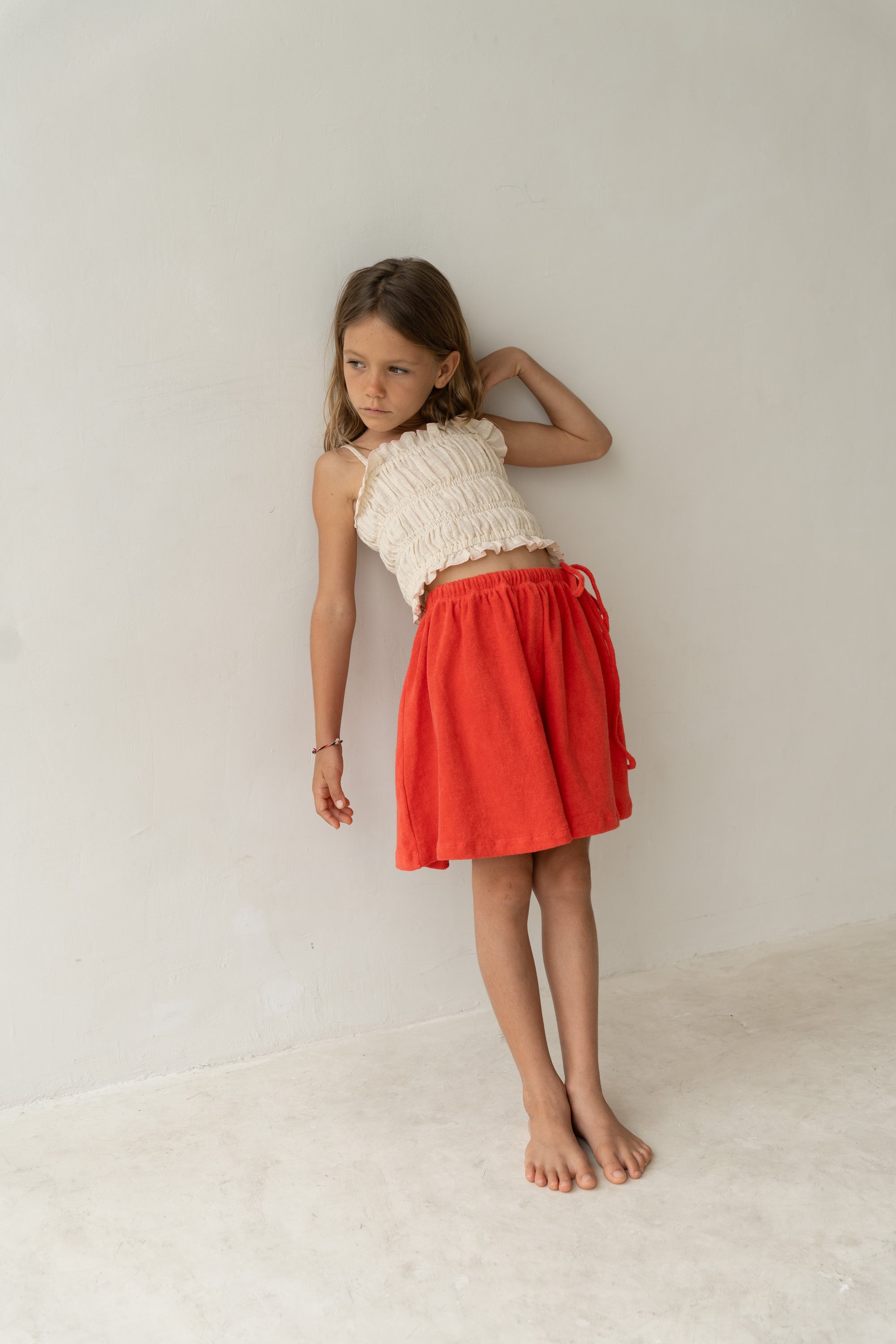 A young girl with long hair stands barefoot, leaning against a wall. She is wearing a white sleeveless top and an ILLOURA THE LABEL Lennie Skirt in watermelon, featuring an elasticated waistband in a vibrant hue. Her expression is thoughtful as she gazes off to the side against the plain, light-colored wall.