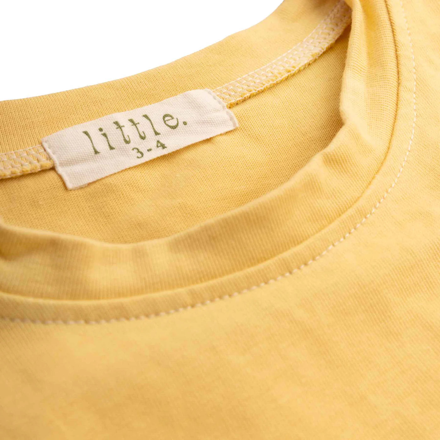 Close-up of a yellow children's tee by LITTLE THE LABEL, "Little Lou Tee Pomello," featuring a white label near the neckline with the word "little" and green text showing size "3-4," emphasizing its 100% cotton comfort.