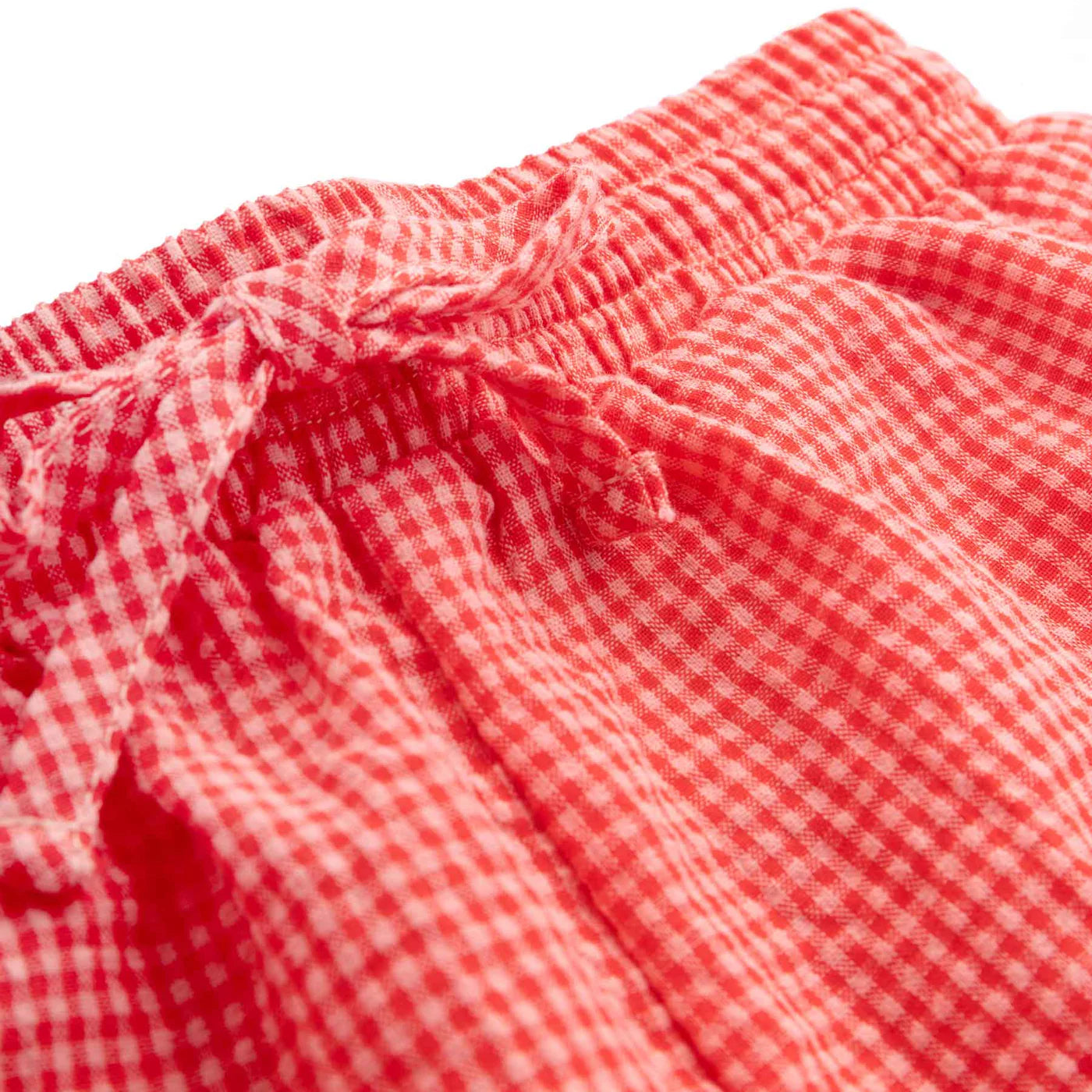 Close-up of red and white cherry gingham fabric, likely part of a garment by LITTLE THE LABEL. The seersucker cotton is gathered with a drawstring, suggesting it might be the waistband for the unisex Cami Shorts Cherry Gingham.