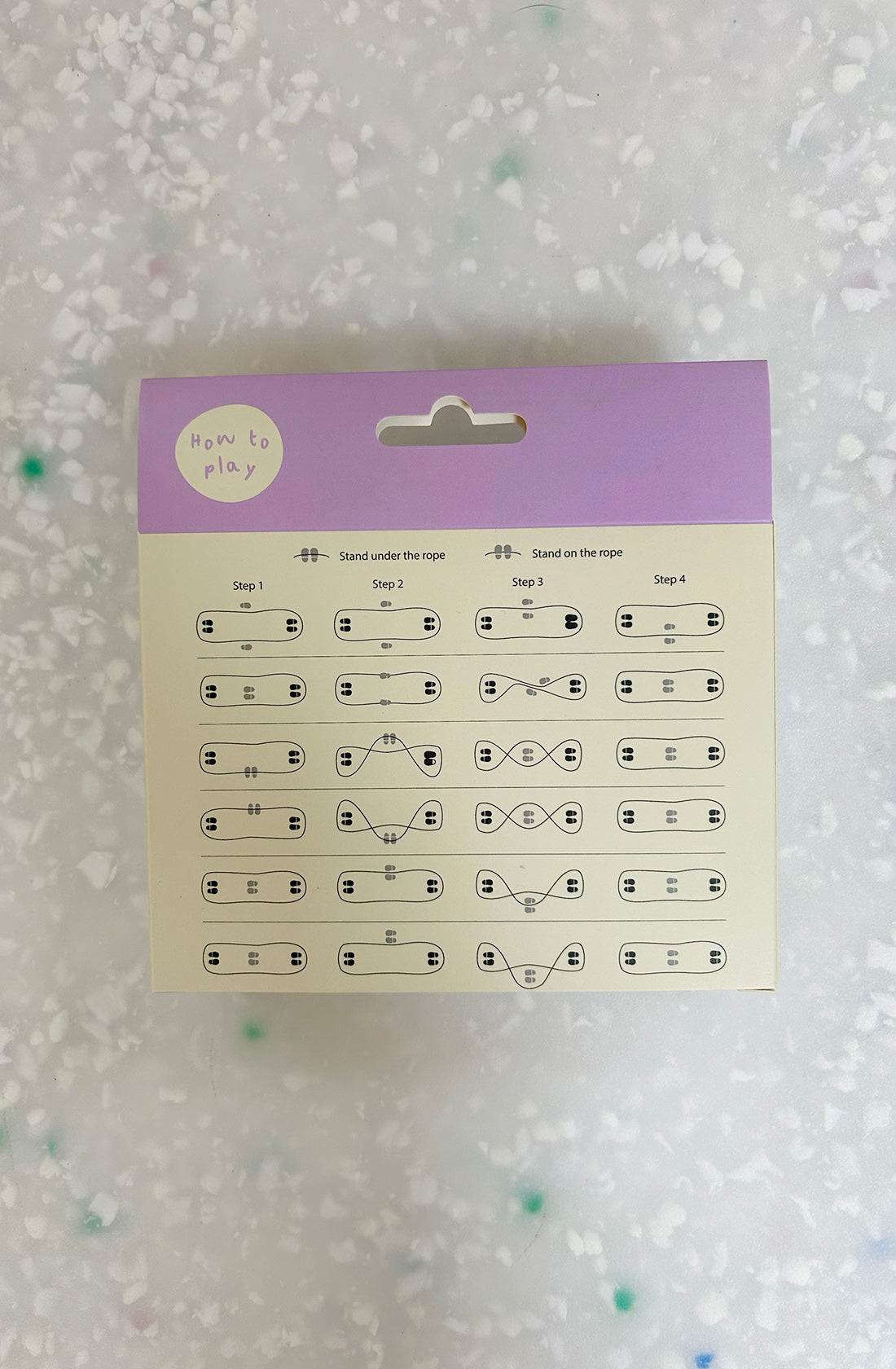 A card with a purple top section labeled "How to play" from FABELAB's Skipping Elastic Lilac set. Below, four steps are illustrated with small icons showing foot placement and movements for outdoor rope play, all on a freckled light surface background.