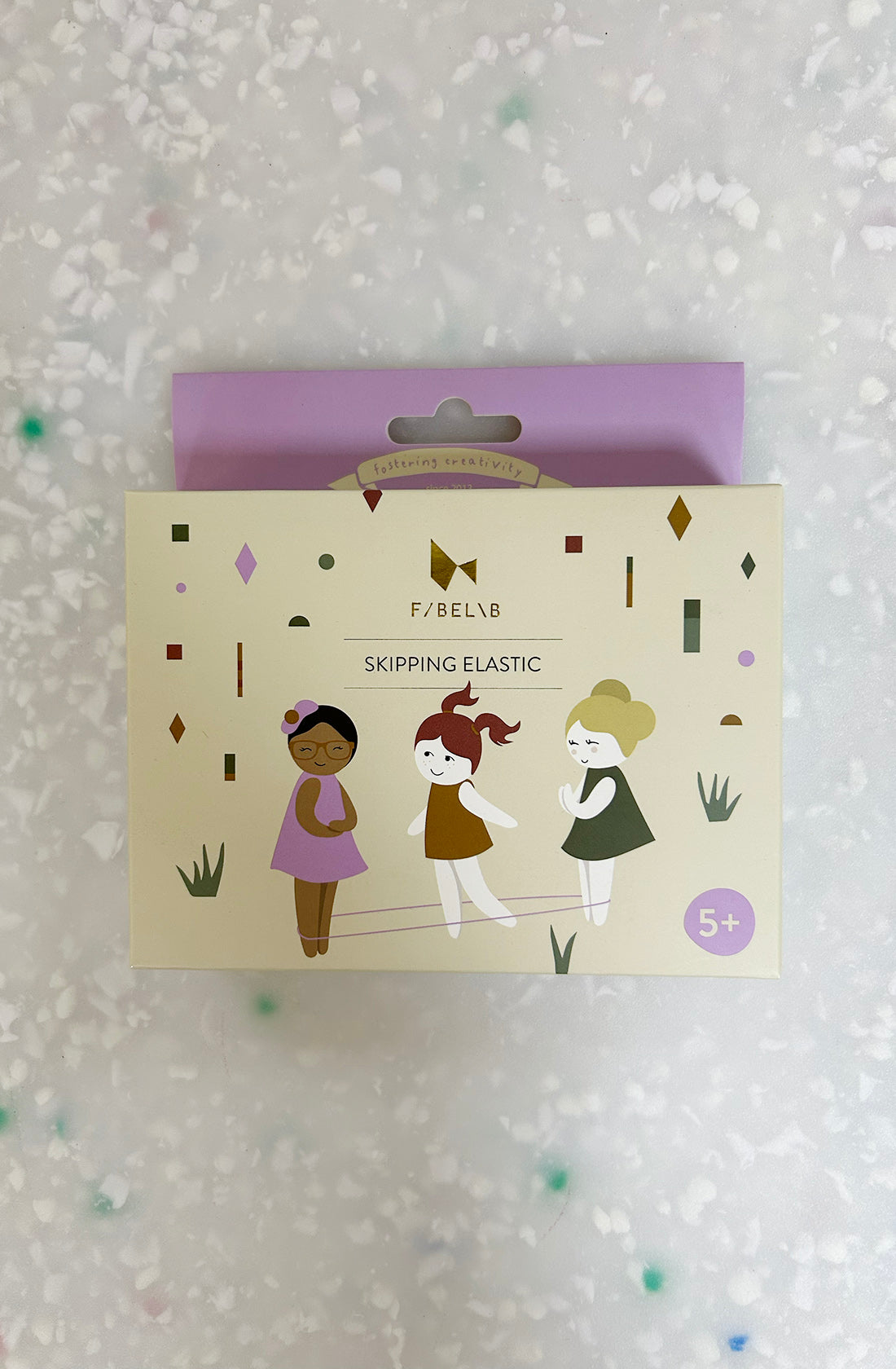 A Skipping Elastic Lilac, part of the summer collection by FABELAB, is packaged in a light-colored box featuring illustrations of three children enjoying outdoor play. One child wearing glasses and a purple shirt holds the elastic, while a red-haired child in a green dress skips and a blonde child in a white dress assists. Suitable for ages 5 and up.