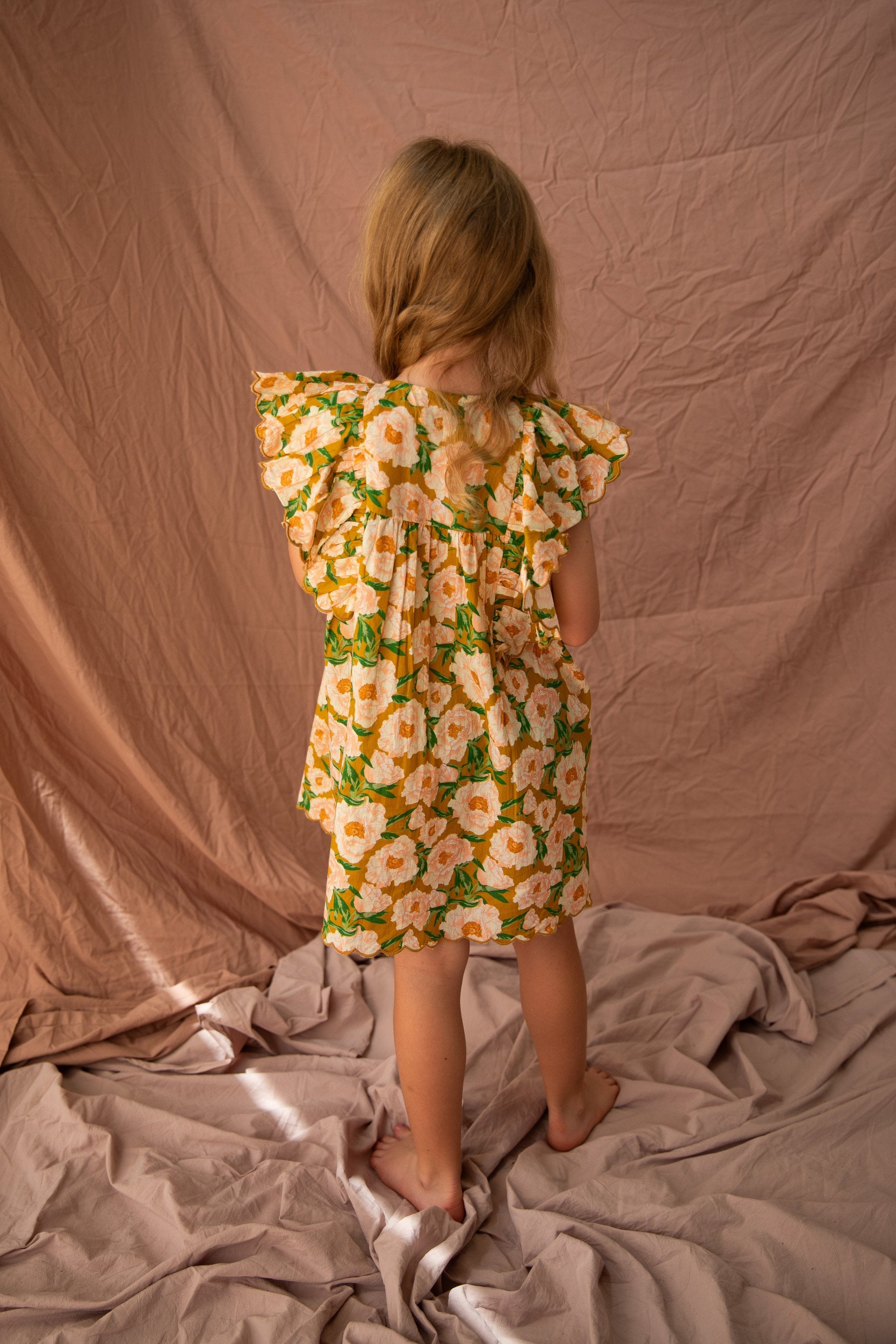 A young child with long blonde hair stands barefoot, facing away from the camera. They are wearing the Sophia Dress in Soirée Bouquet/Honey Mustard by BELLA + LACE, featuring frilled shoulders. The child is standing in front of a backdrop of crumpled pink fabric. The setting appears to be indoors, and the lighting is soft.