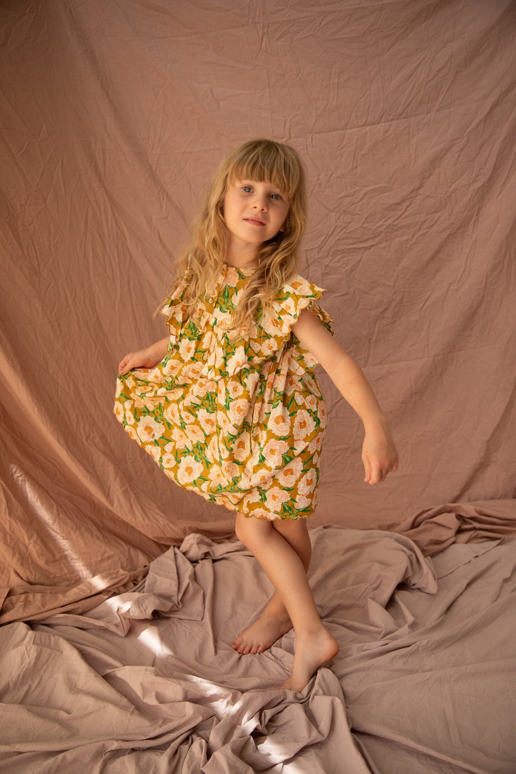 A young girl with long blonde hair dances barefoot on a draped fabric backdrop. She wears the BELLA + LACE Sophia Dress Soirée Bouquet/Honey Mustard, which is adorned with white flowers and frilled shoulders, and her expression is serene. The neutral beige color of the backdrop softly contrasts with her vibrant dress.