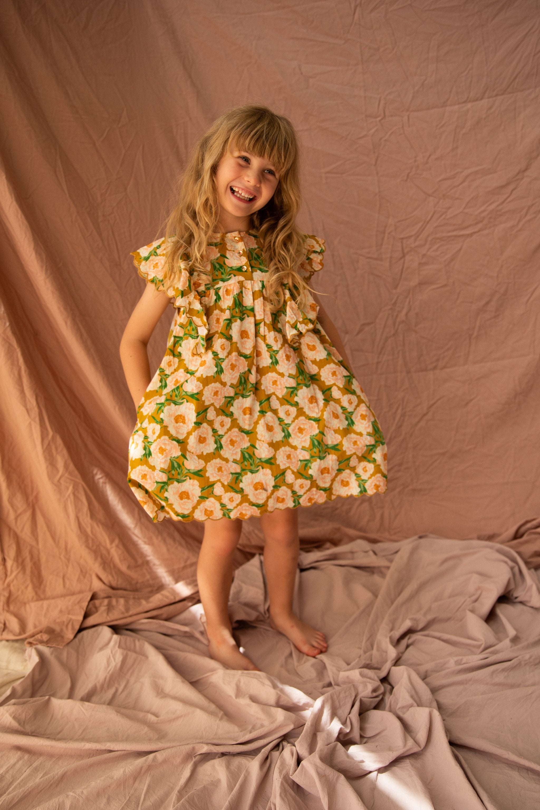 A young girl with long blonde hair stands barefoot on wrinkled fabric. She is smiling and wearing the Sophia Dress Soirée Bouquet/Honey Mustard by BELLA + LACE, featuring frilled shoulders adorned with yellow and white flowers with green leaves. The backdrop is a mix of light brown and beige cloth.