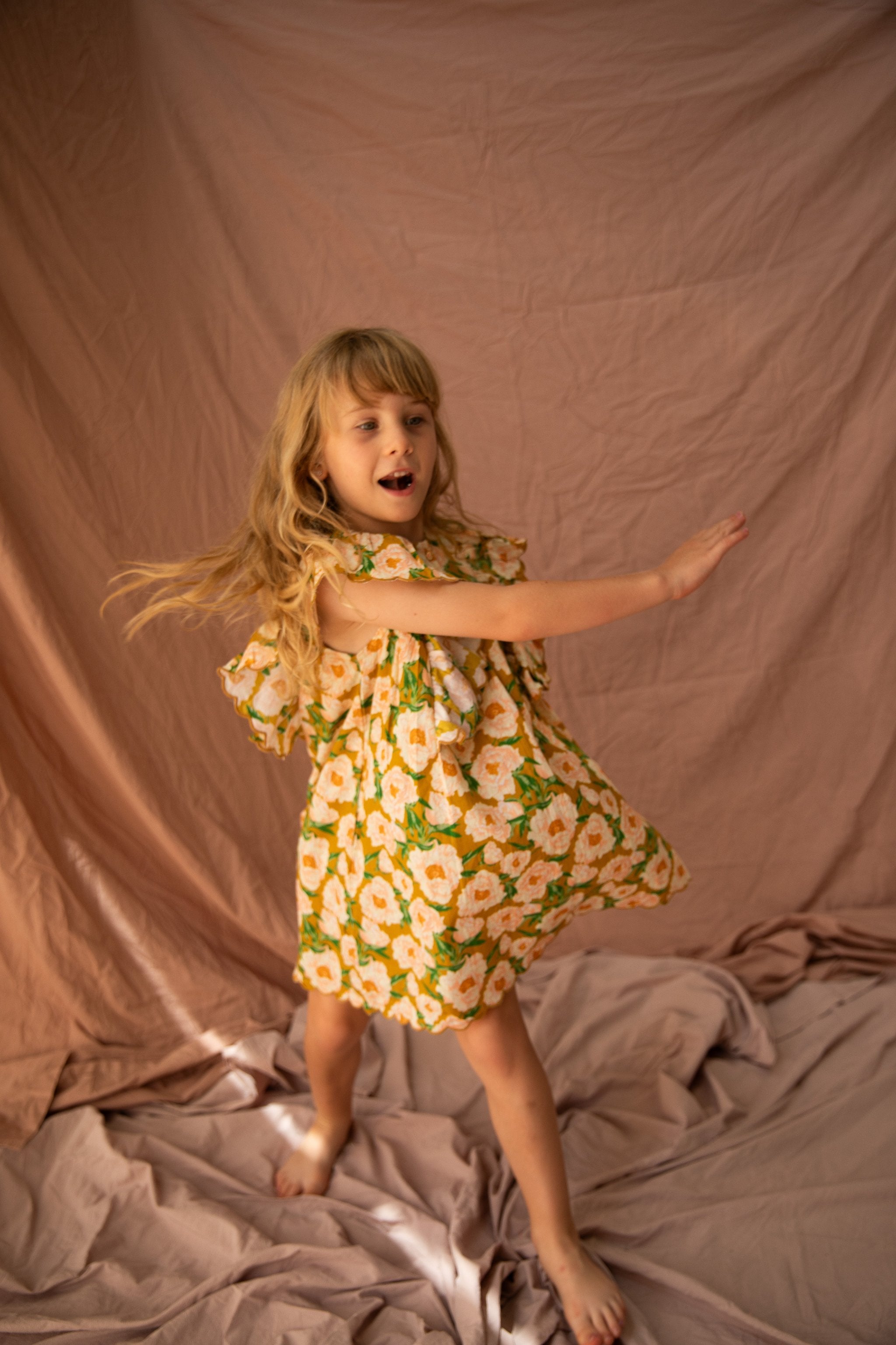 A young girl with long blond hair dances joyfully in the BELLA + LACE Sophia Dress Soiree Bouquet/Honey Mustard, which is adorned with a beautiful Soirée Bouquet print and frilled shoulders. The background is draped with soft, light brown fabric, creating a warm and playful atmosphere. Her arms are outstretched, and her dress flows as she moves.