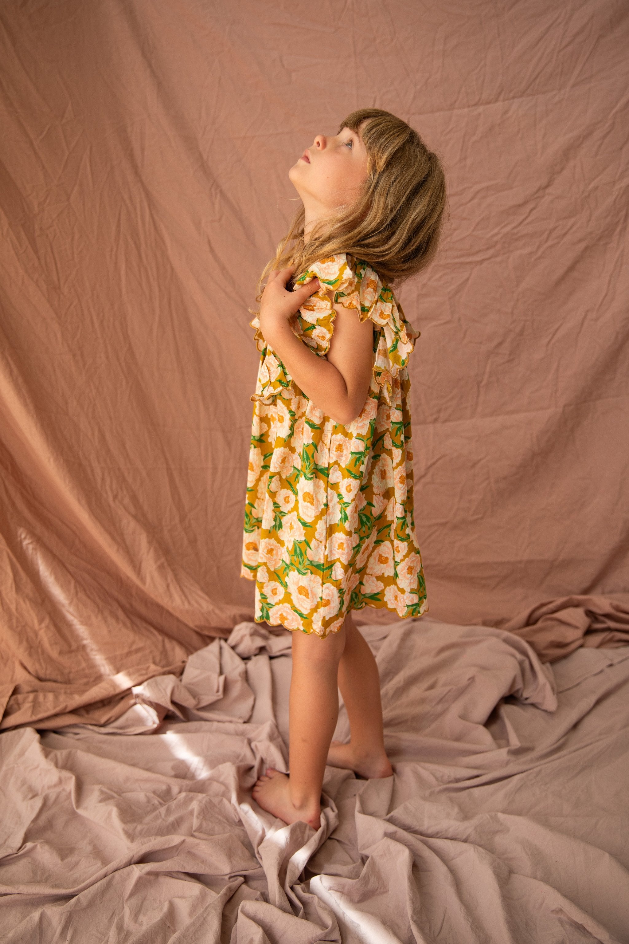 A young girl wearing the BELLA + LACE Sophia Dress in Soirée Bouquet/Honey Mustard stands barefoot on a crumpled fabric, looking upwards with one hand touching her chest and the other by her side. The backdrop is a slightly wrinkled, soft pink fabric.