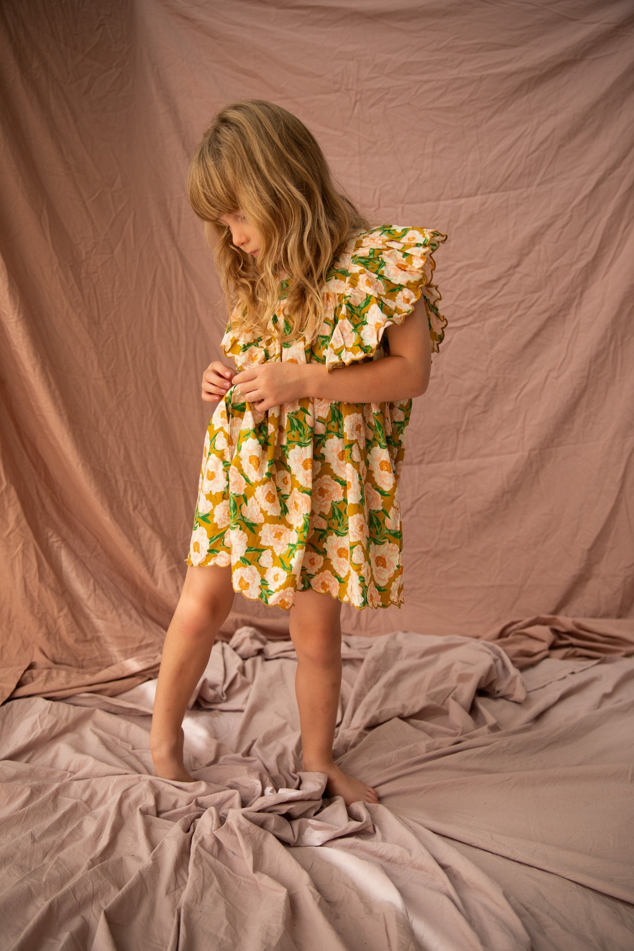 A young girl with long, blonde hair stands barefoot on a floor covered with crumpled, light brown fabric. She wears the Sophia Dress Soirée Bouquet/Honey Mustard by BELLA + LACE, featuring frilled shoulders and decorated in a yellow floral pattern. She looks down, gently lifting her dress with her hands. The background is draped with matching fabric.