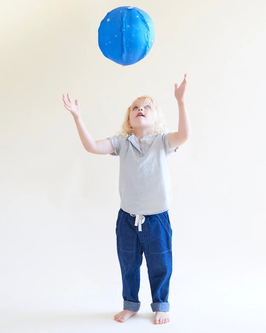 A young child with light hair playfully tosses the Starry Night Balloon Ball by SARAH'S SILKS, which is adorned with yellow stars. The barefoot child, dressed in a gray shirt and blue pants, looks up at the ball against a plain, light background.