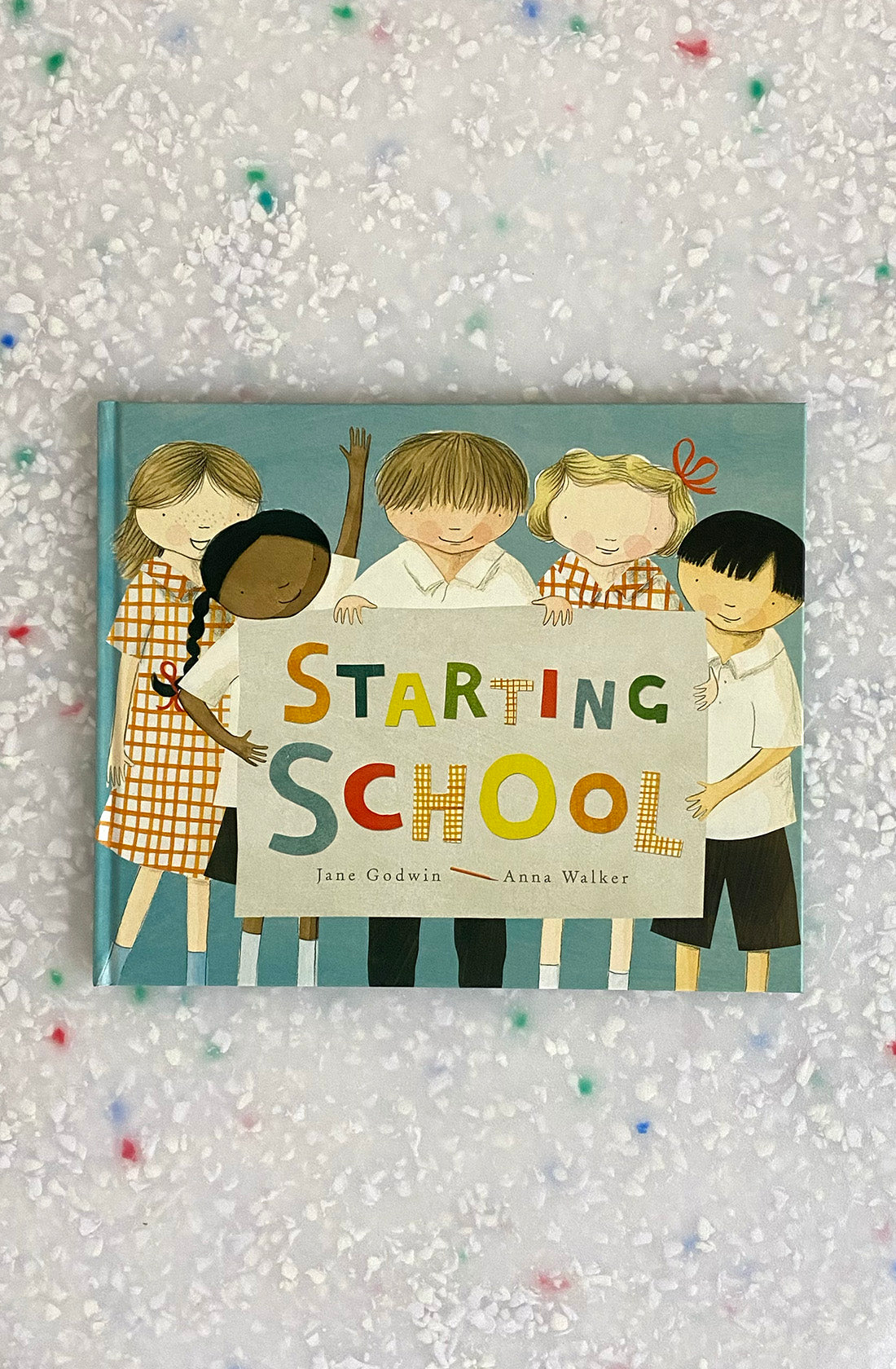 Cover of the children's book "Starting School" by Jane Godwin and Anna Walker, published by BAY KIDS. The illustration features five children holding up a sign with the book title, making it ideal for the first day of school. The background is a speckled surface, and each letter in the title is in a different color.