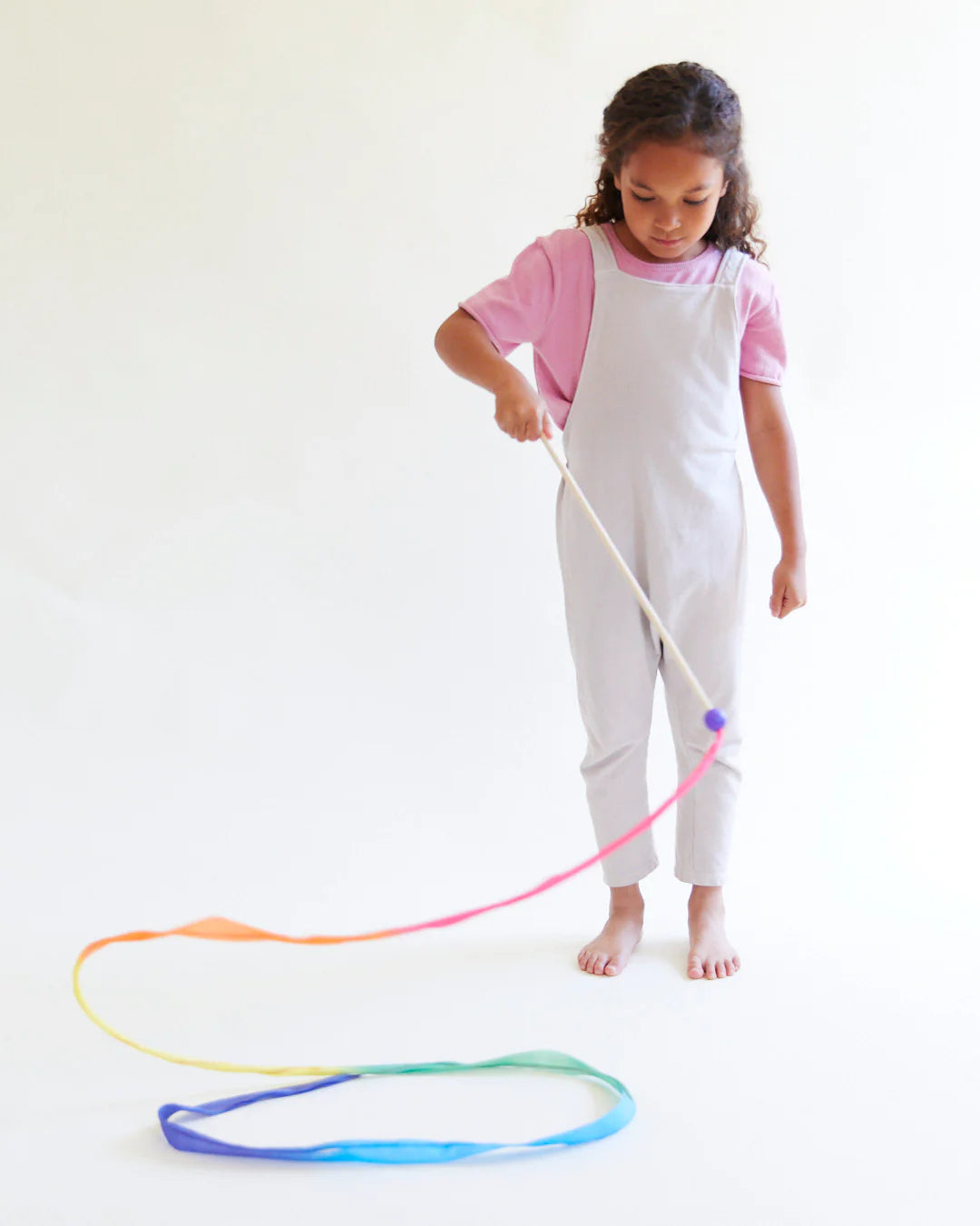 A young girl dressed in a pink shirt and white overalls is playing with the Large Rainbow Streamer from SARAH'S SILKS, crafting flowing shapes on the floor. Holding a wooden wand crafted from sustainably sourced materials, her imagination dances against the plain white background.