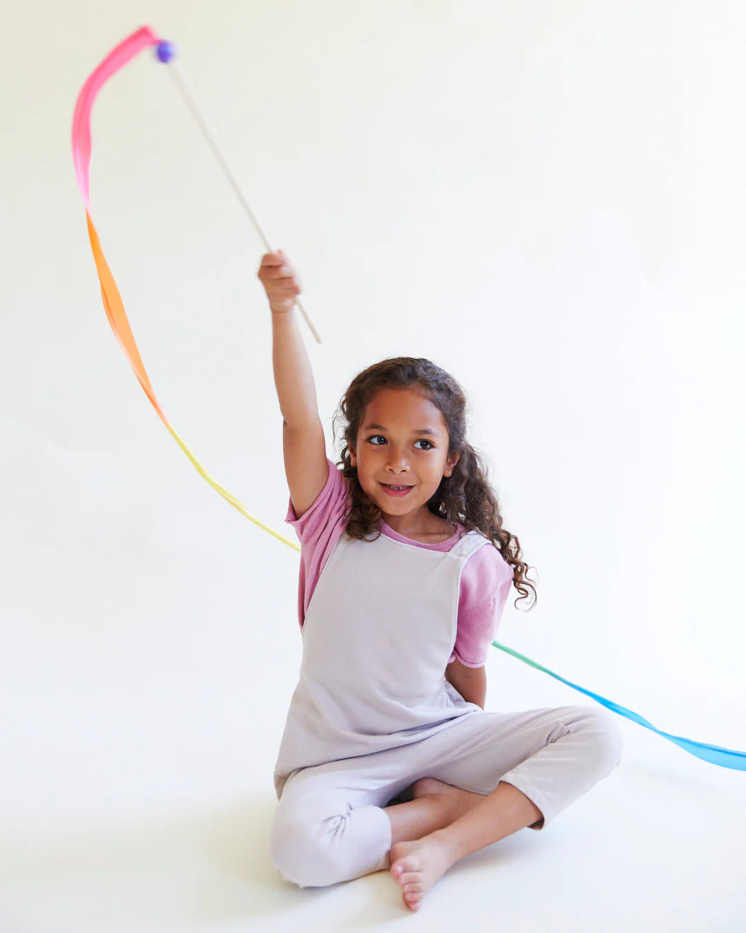 A young girl sits cross-legged on the floor, holding SARAH'S SILKS' Large Rainbow Streamer. She wears a pink shirt under a light grey dress and looks joyful against a plain background.