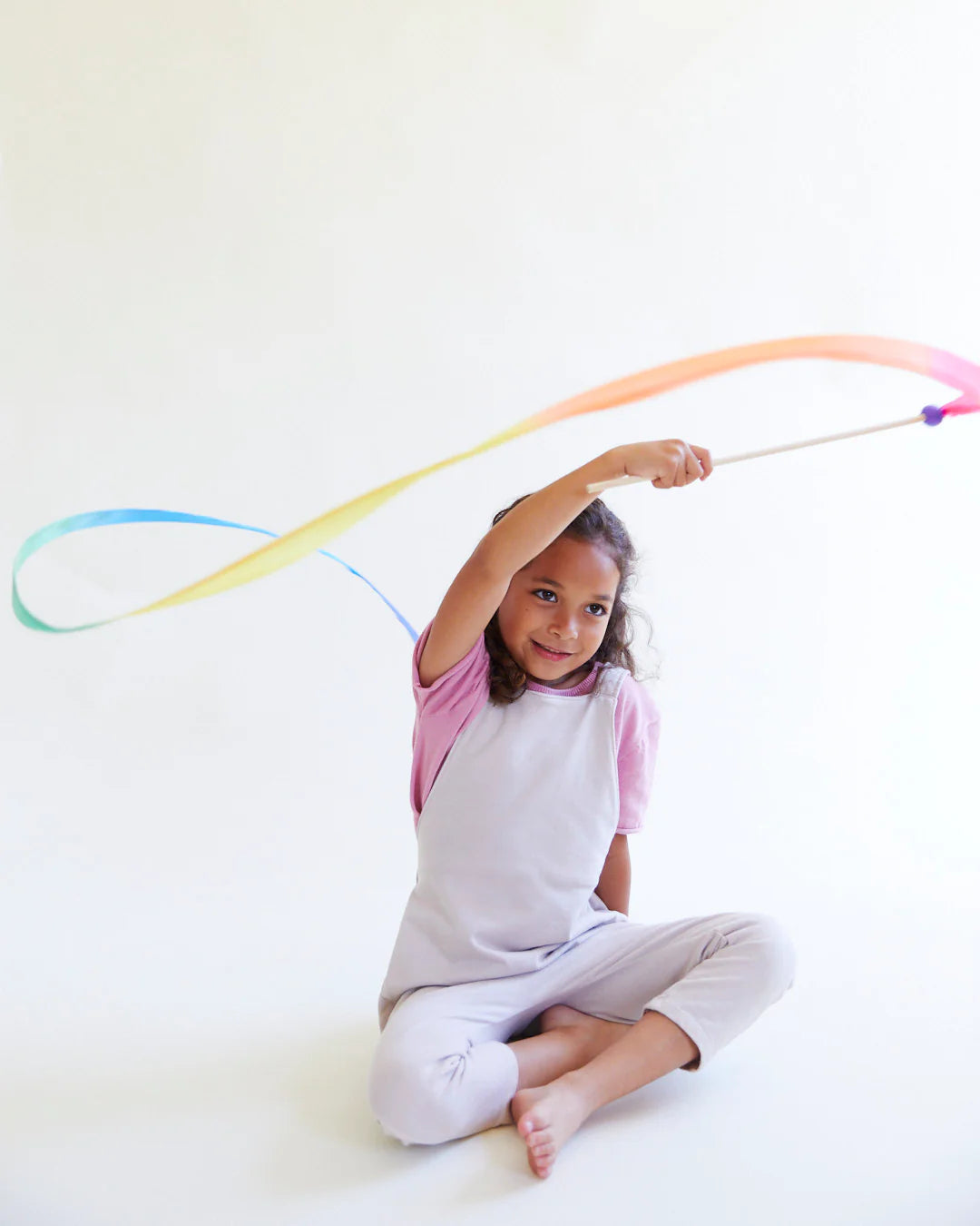 A child sits on the floor, gracefully waving a Large Rainbow Streamer from SARAH'S SILKS with a wooden wand, forming an elegant arc. Clad in a light pink t-shirt and white overalls, the scene unfolds against a plain white background.