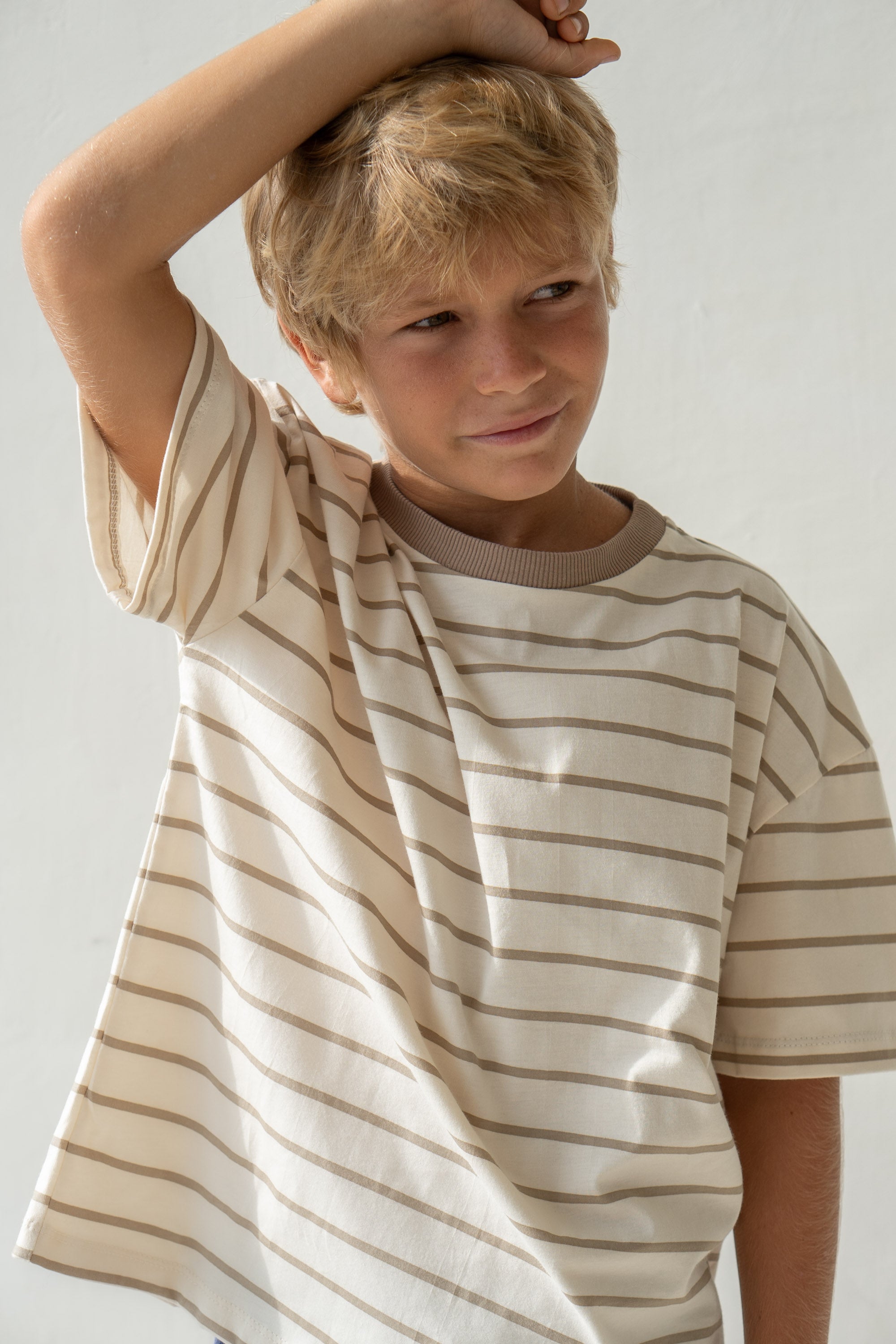 A young boy with blond hair smiles subtly as he wears the Basic Tee Mushroom Stripe by ILLOURA THE LABEL, crafted from breathable cotton. He has one arm raised, resting on his head against a neutral background.