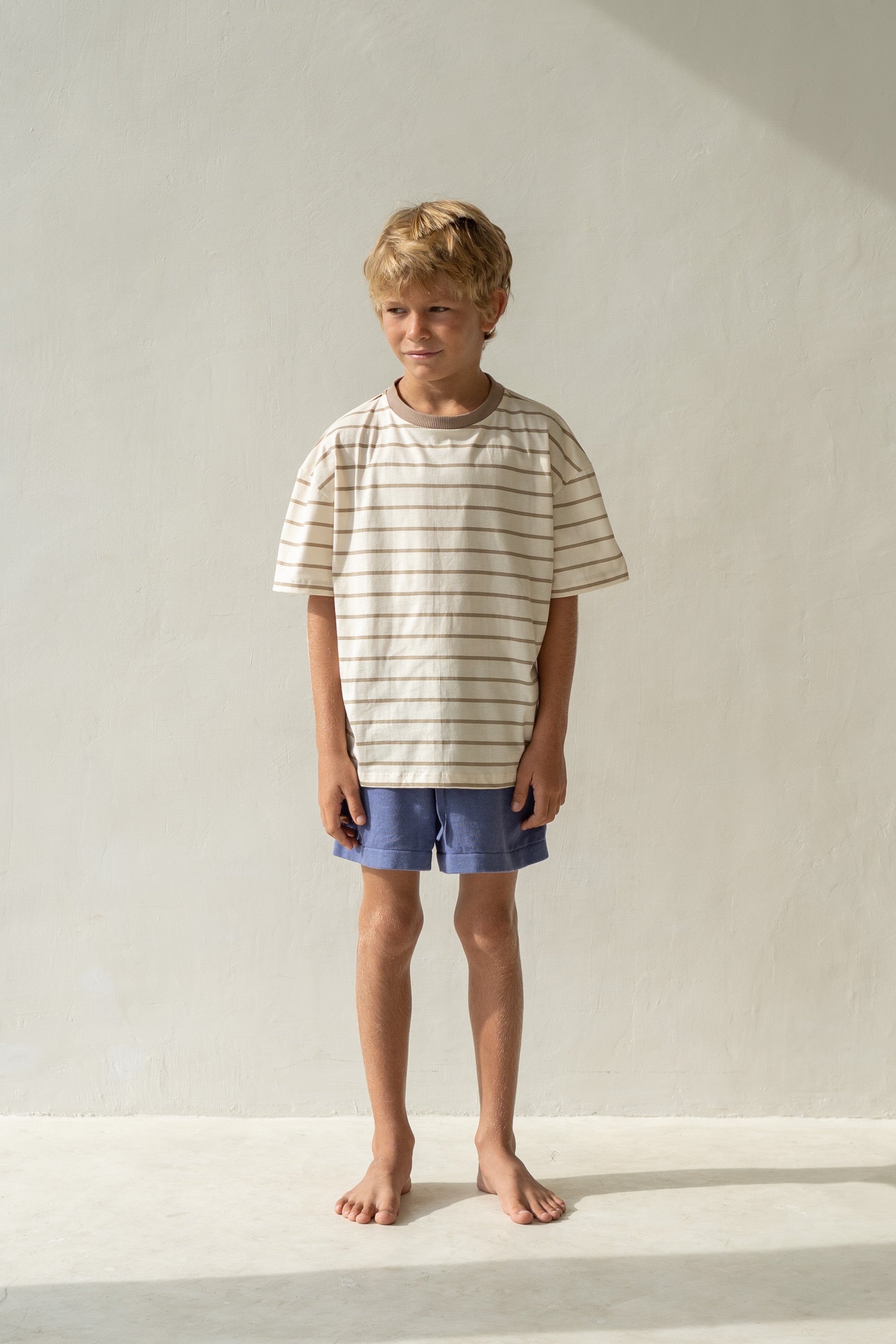 A young boy with blonde hair stands barefoot on a light-colored floor, dressed in an ILLOURA THE LABEL Basic Tee Mushroom Stripe made from breathable cotton and blue shorts, looking slightly to the side. The wall behind him is plain and light-colored.