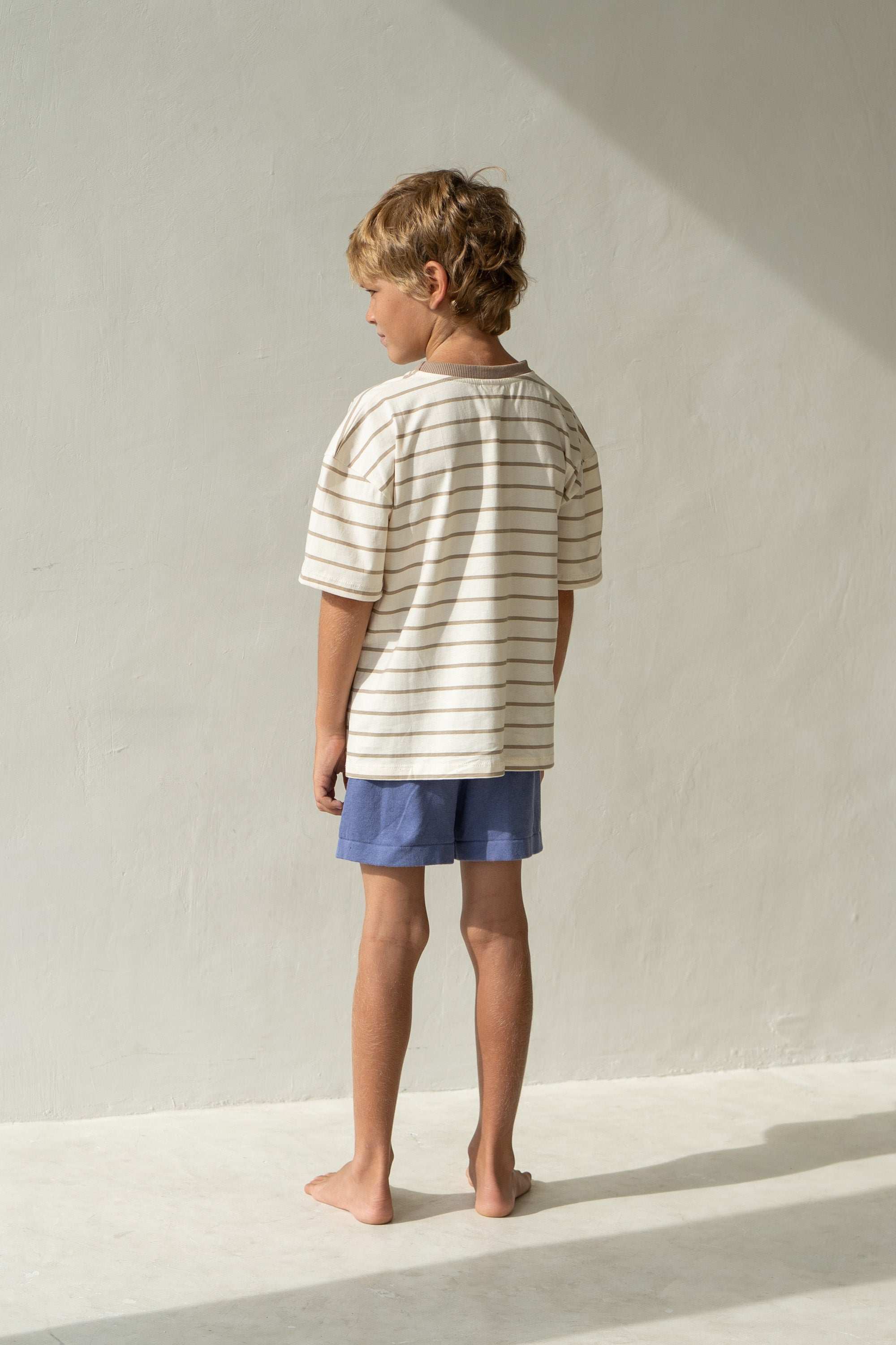 In a softly lit room, a child with curly hair stands barefoot, facing away. They are wearing the "Basic Tee Mushroom Stripe" by ILLOURA THE LABEL paired with blue shorts. The light-colored wall and floor are gently illuminated by sunlight, casting a delicate shadow.