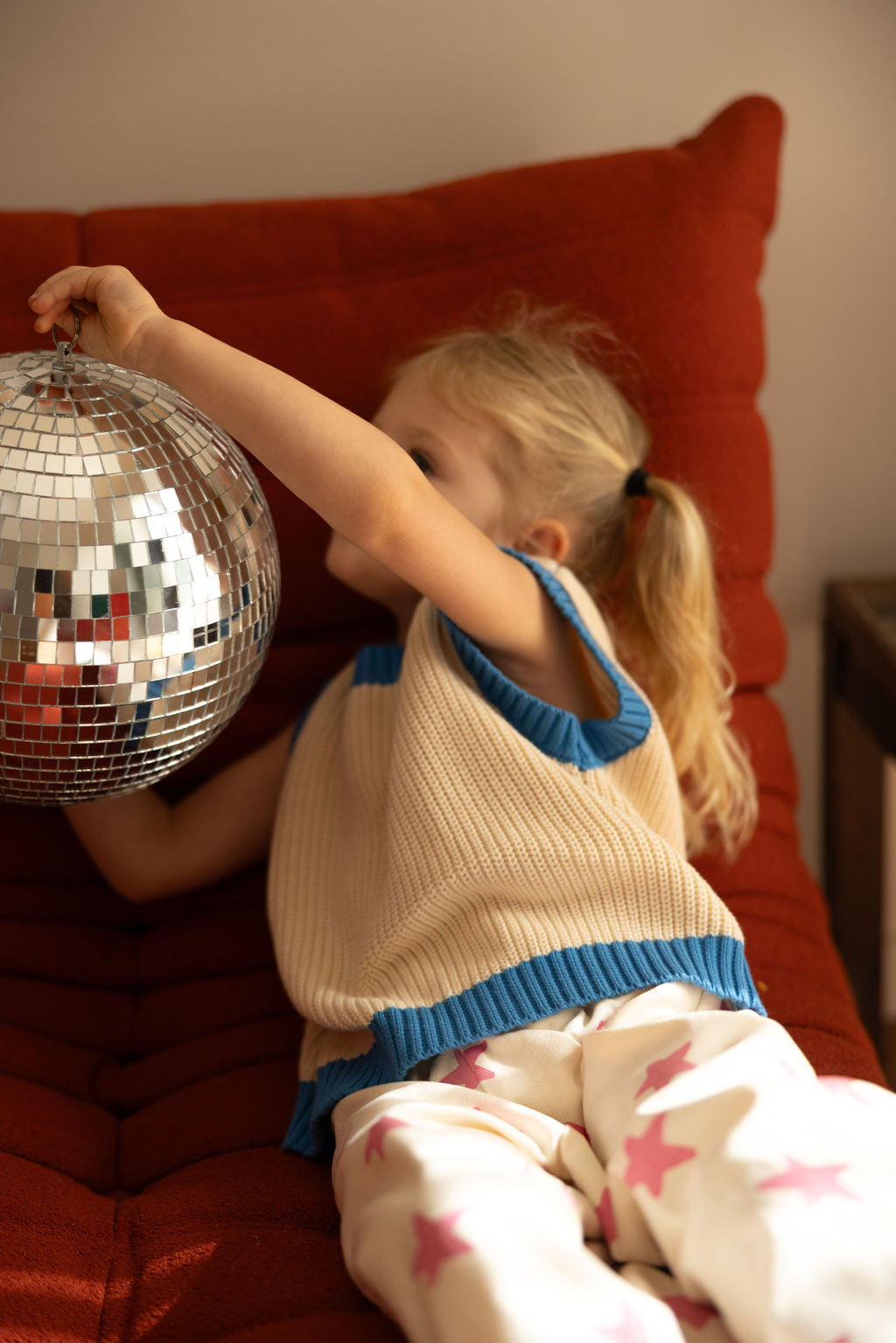 A child with blonde hair, wearing SUNDAY SIBLINGS' Nonno Vest Blue and white pants embellished with pink stars, lies on a red sofa. They hold a shiny disco ball in both hands in the softly lit room.