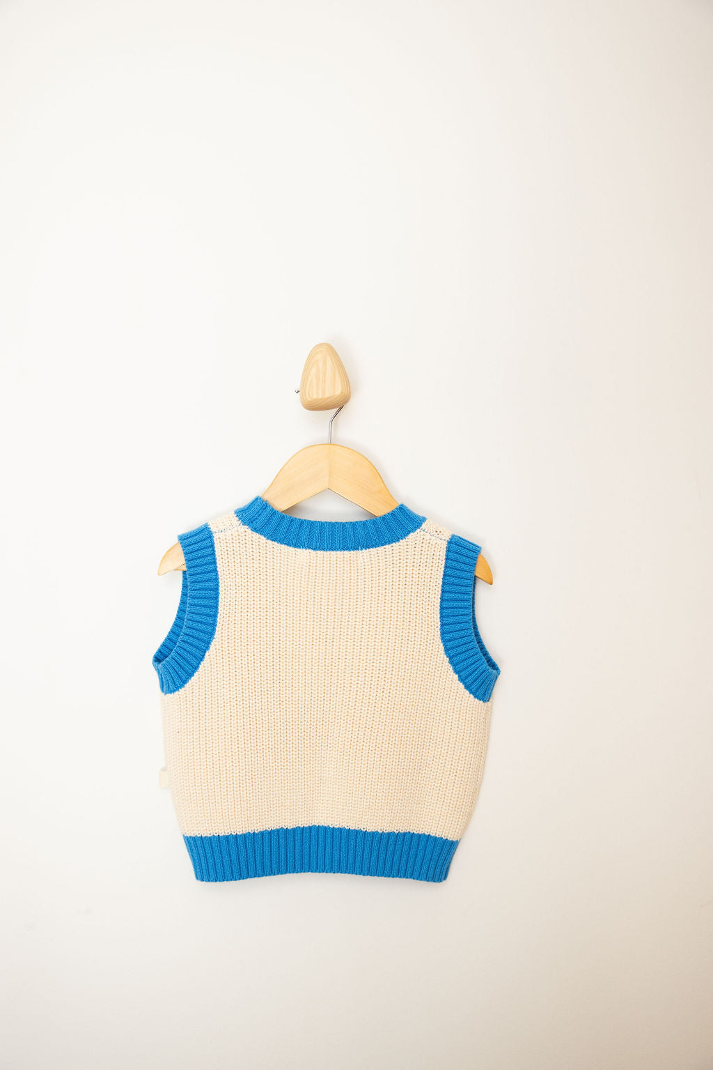 A cream-colored knitted SUNDAY SIBLINGS Nonno Vest Blue, featuring a blue trim and offering a charming grandad aesthetic, is displayed on a wooden hanger against a plain white wall. Crafted from 100% cotton, it ensures both comfort and style.