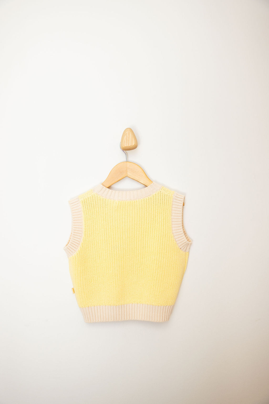 The "Nonno Vest Butter Yellow" by SUNDAY SIBLINGS, a sleeveless knit top made from 100% cotton with cream-colored trim, elegantly hangs on a wooden hanger against a plain white background.