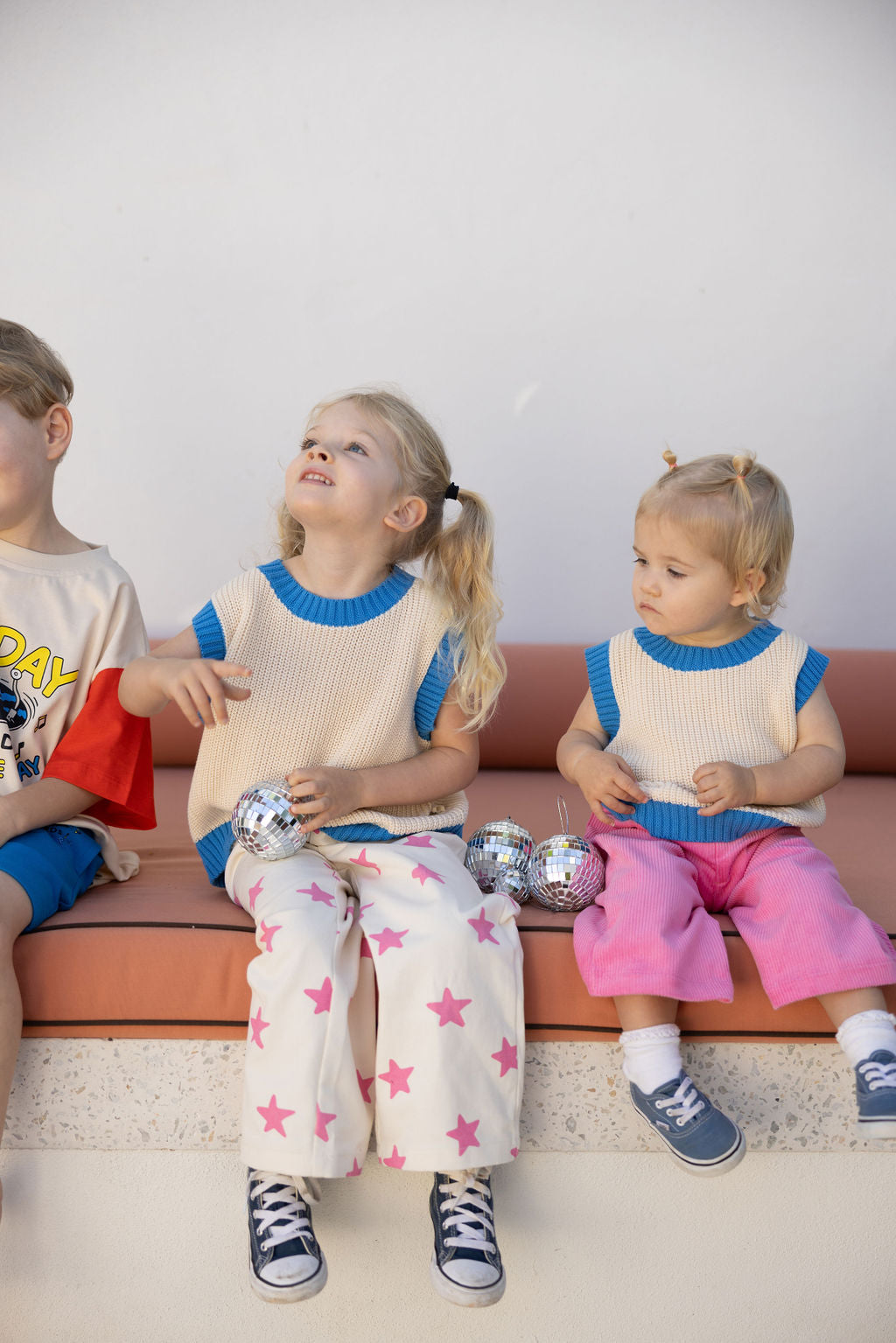 Two young children sit on a bench holding small disco balls. The child on the left, with light blonde hair in pigtails, wears a sleeveless Nonno Vest Blue by SUNDAY SIBLINGS and star-patterned pants. The child on the right wears a matching sweater made from 100% cotton and pink pants, looking to the side.