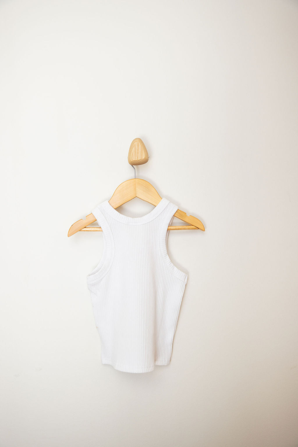 The PRE-ORDER Minnie Singlet from SUNDAY SIBLINGS, a white sleeveless ribbed cotton top, is displayed on a wooden hanger against a plain off-white wall, subtly echoing sophistication with its embroidered logo.