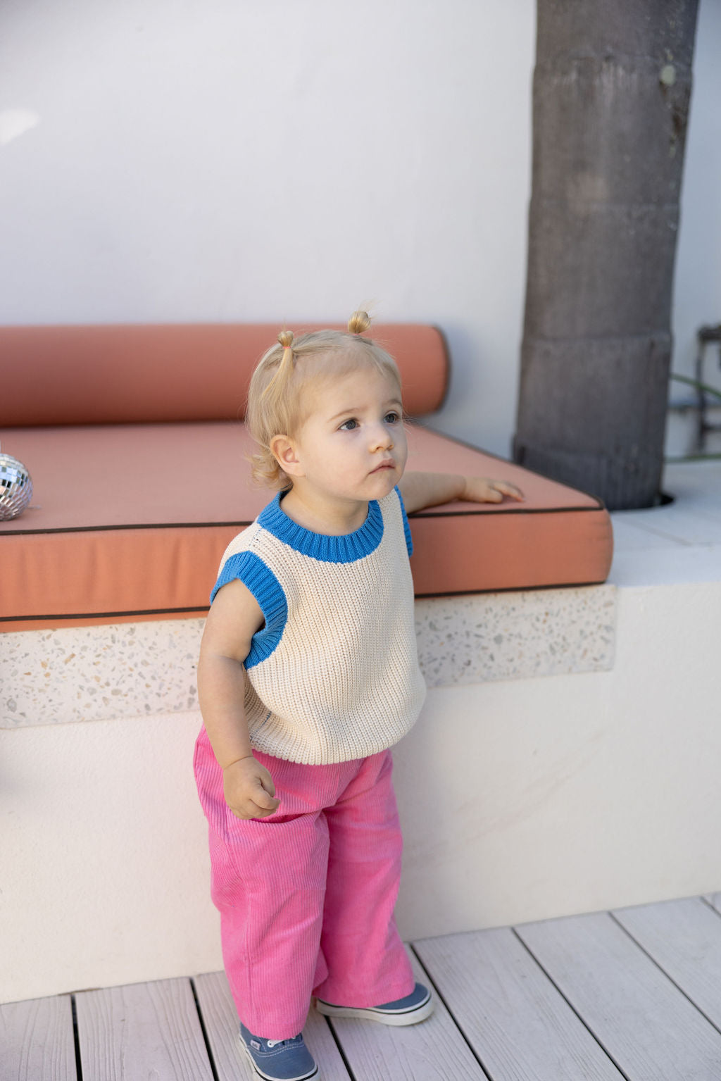 A toddler with two small ponytails stands beside a cushioned bench, looking curious and attentive while resting one hand on it. The child is wearing the cream-colored Nonno Vest Blue by SUNDAY SIBLINGS over a blue top, paired with pink pants and blue shoes.