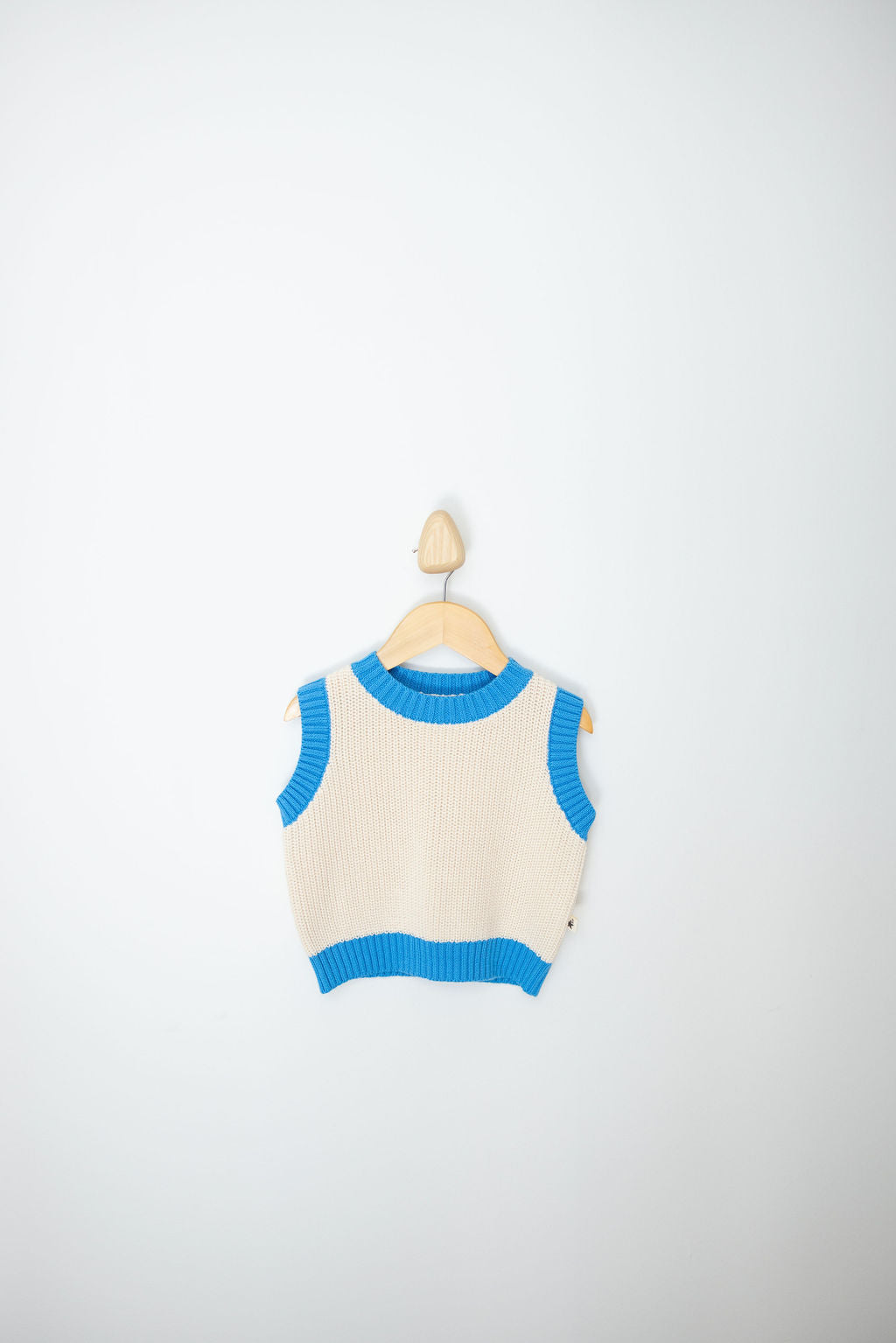 A sleeveless Nonno Vest Blue from SUNDAY SIBLINGS is displayed on a wooden hanger against a plain background. Made from 100% cotton, this cream-colored vest offers a touch of grandad style with its bright blue accents on the collar, armholes, and hem. The minimalist setup emphasizes the vest's design and contrasting colors.