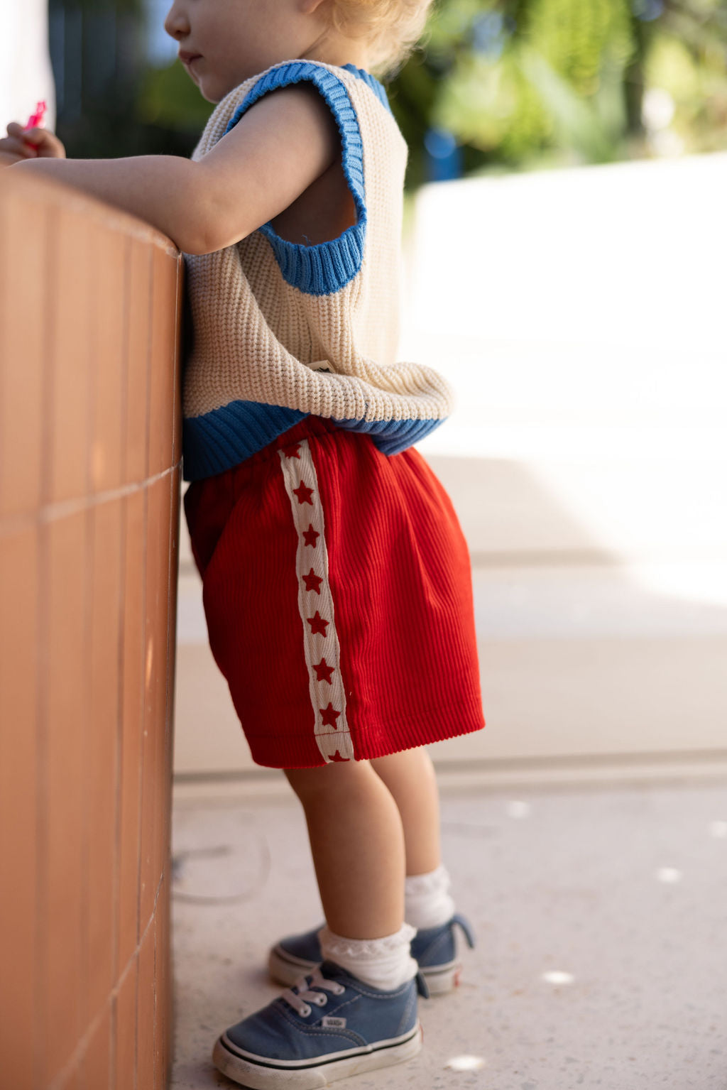 A toddler dressed in a sleeveless knit top and SUNDAY SIBLINGS' Playtime Cord Shorts in red, featuring star embroidery, stands next to a low wall. The child wears blue sneakers and white socks, while sunlight casts a soft glow on the scene.