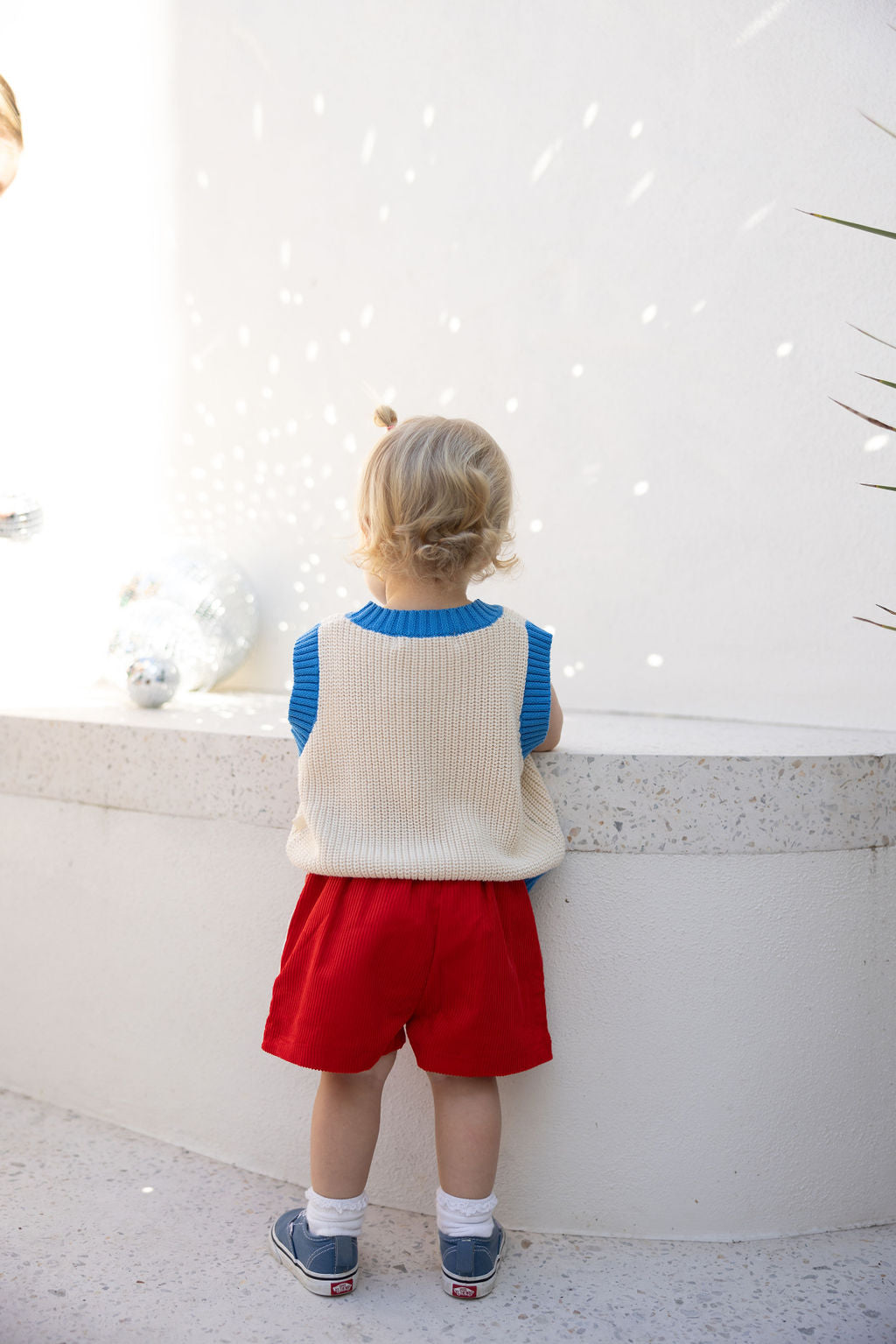 A toddler with light hair faces away from a white wall. They're dressed in a blue shirt, cream vest, and vibrant red Playtime Cord Shorts Red from SUNDAY SIBLINGS, featuring an elastic waistband. Sneakers complete the ensemble as light reflections form patterns on the wall.