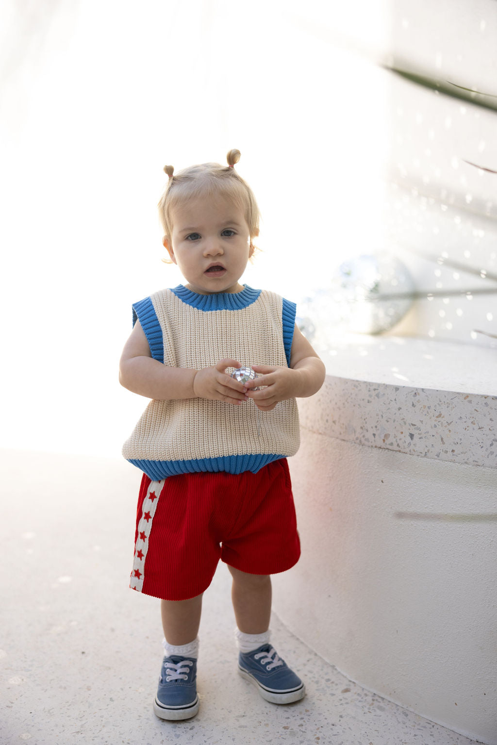 A toddler wearing a cream and blue sweater, SUNDAY SIBLINGS Playtime Cord Shorts in red with star embroidery, and blue shoes stands indoors holding a shiny object. The child's hair is styled in two small buns. White and light reflections cast on the wall enhance the playful ambiance.