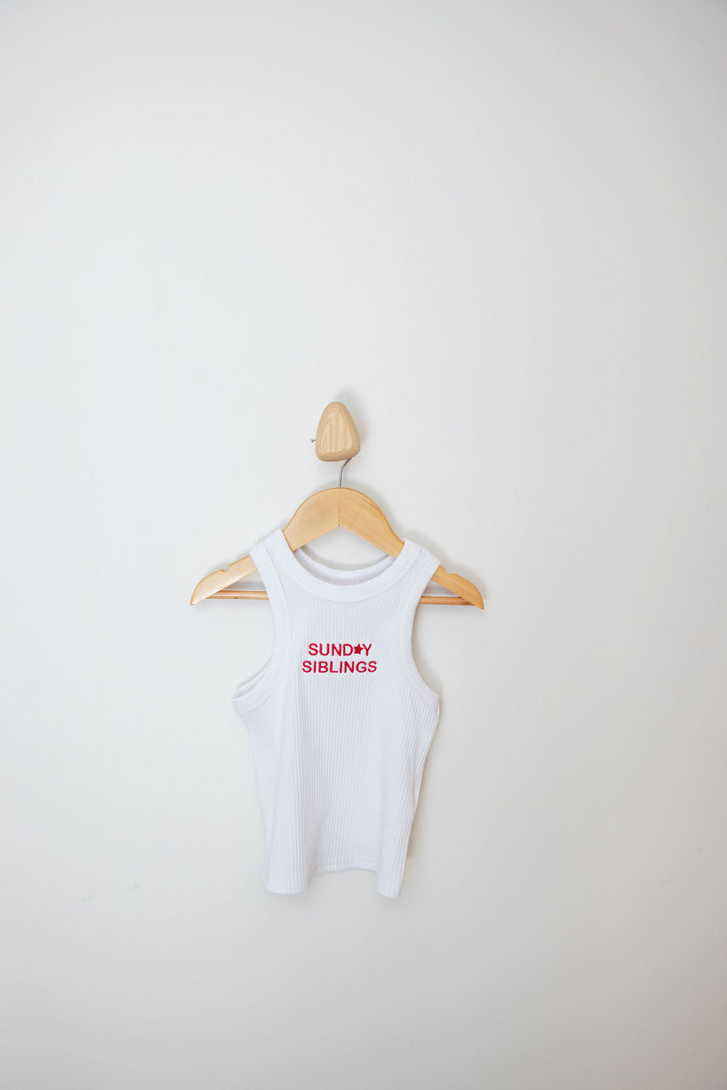 A wooden hanger displays the PRE-ORDER Minnie Singlet, a white sleeveless tank top by SUNDAY SIBLINGS, made from ribbed cotton fabric with "SUNDAY SIBLINGS" printed in red, against a plain white wall.